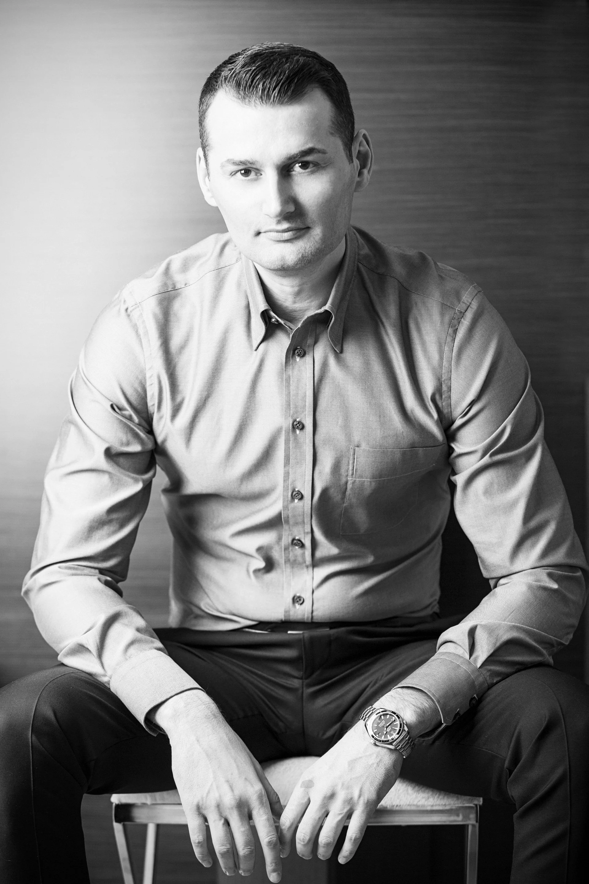 Black and white portrait of a young man sitting on a chair, wearing a button-up shirt and a wristwatch, with a plain background.