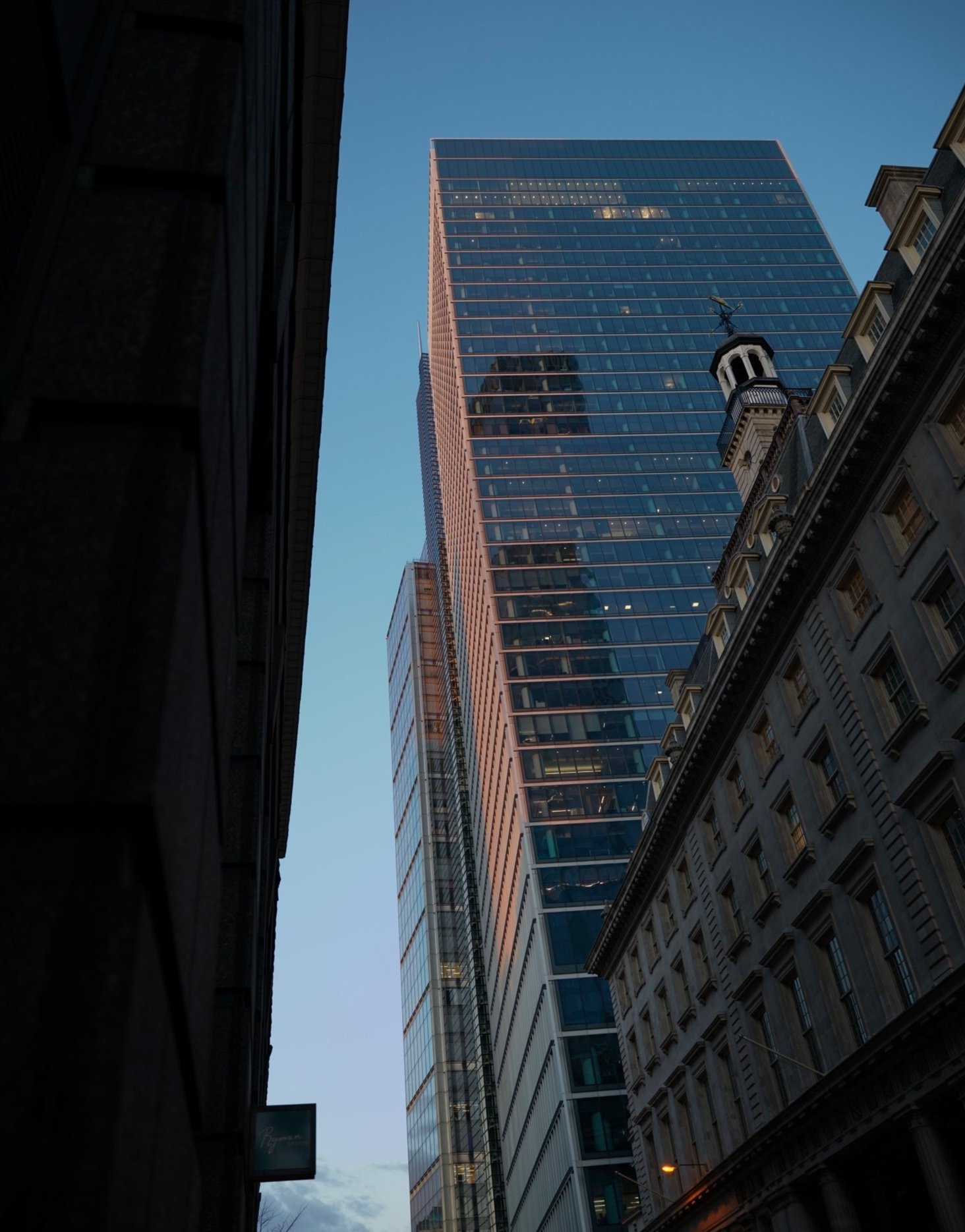 Tall modern glass skyscraper towering above older buildings on a city street, with a clear blue sky in the background.