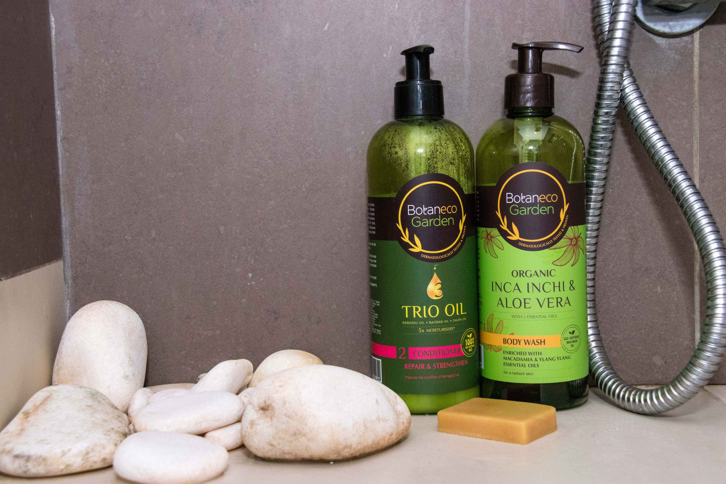 Two green bottles of Botaneco Garden personal care products, a bar of soap, white stones, and a shower hose on a bathroom shelf.