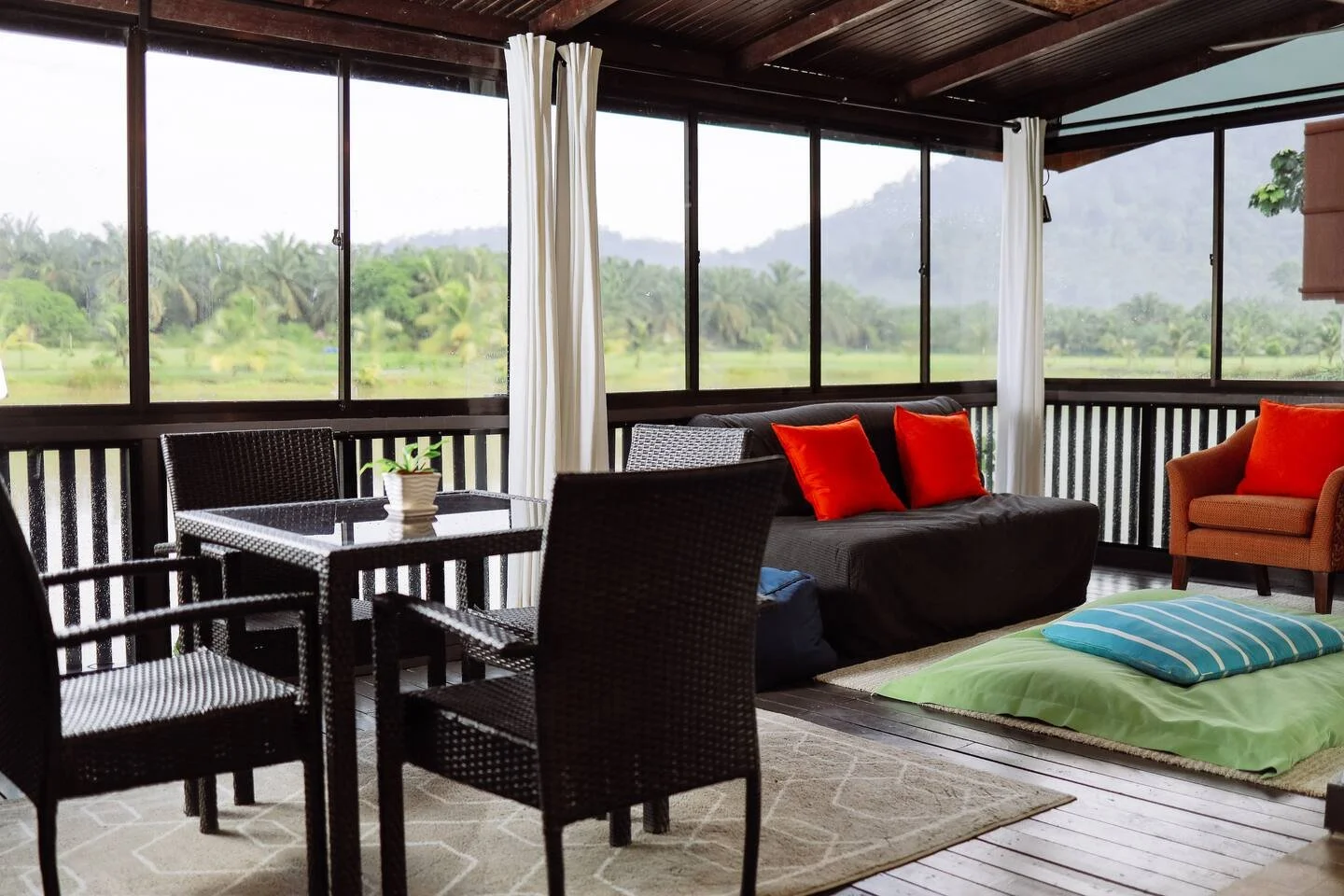 A cozy enclosed porch with large windows overlooking a lush green landscape. There is a small table with four wicker chairs, a dark sofa with red pillows, an orange armchair with a red pillow, and a floor cushion with a blue and white striped pillow 