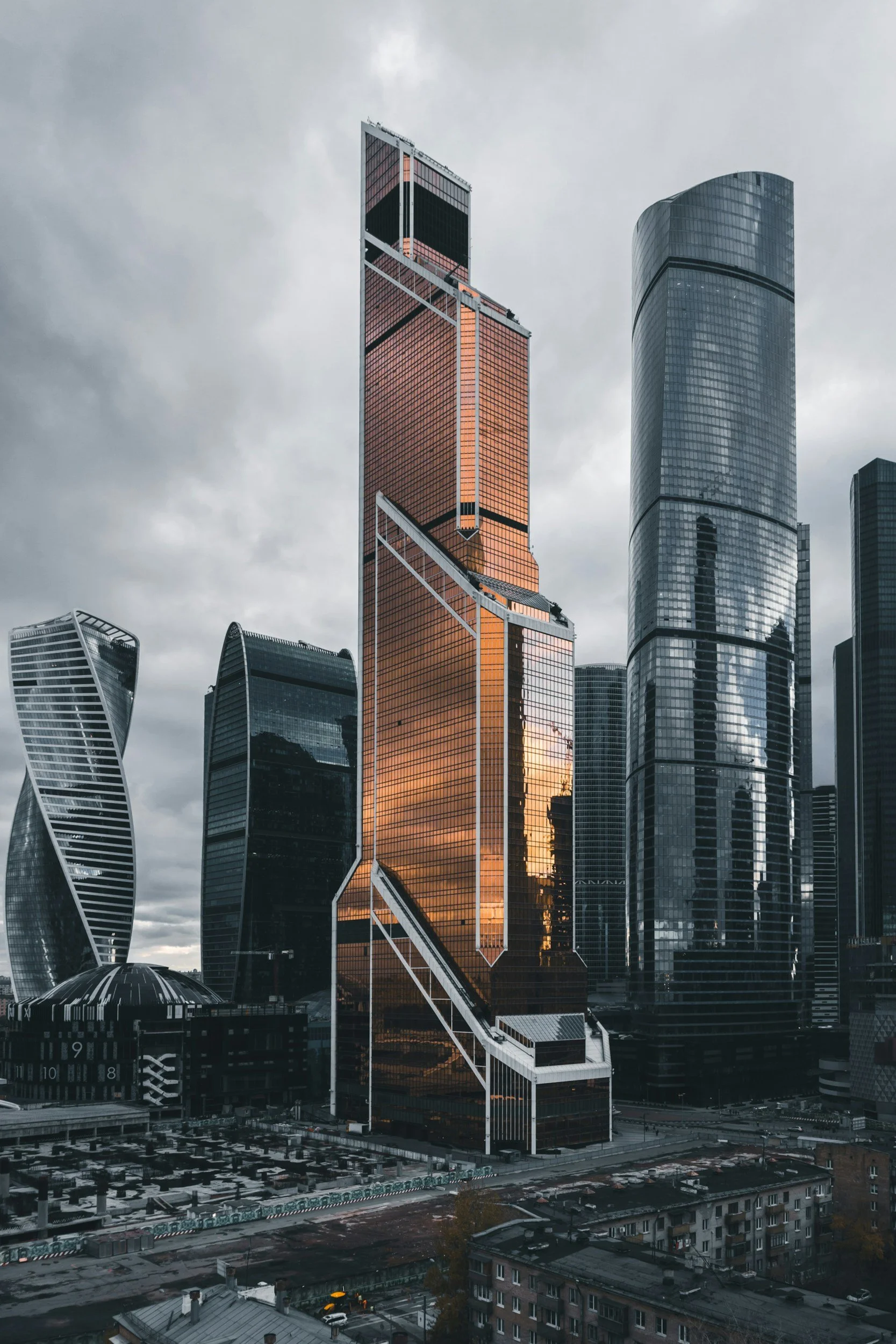 Cityscape featuring modern skyscrapers, including a tall reddish-brown glass building with geometric white accents, overcast sky, and surrounding high-rise buildings reflecting the cloudy weather.