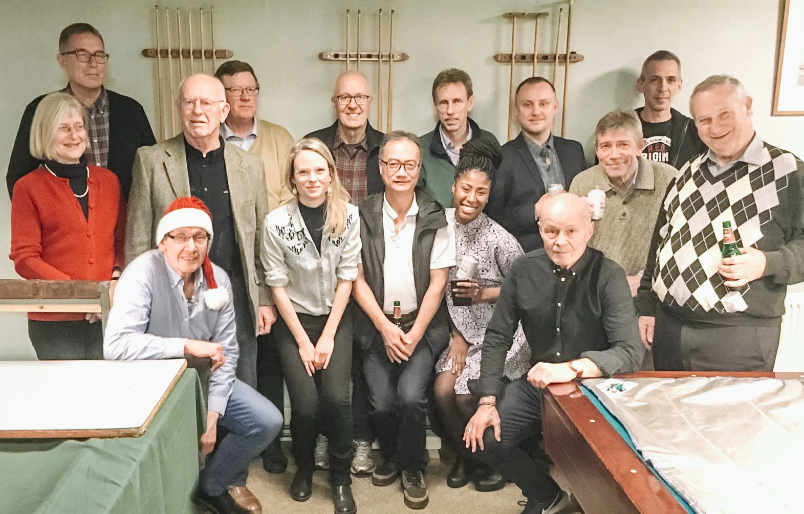 En gruppe mennesker, der fejrer julefrokost, samlet for at tage et foto. Nogle holder drinks, og en person har en nissehue på.