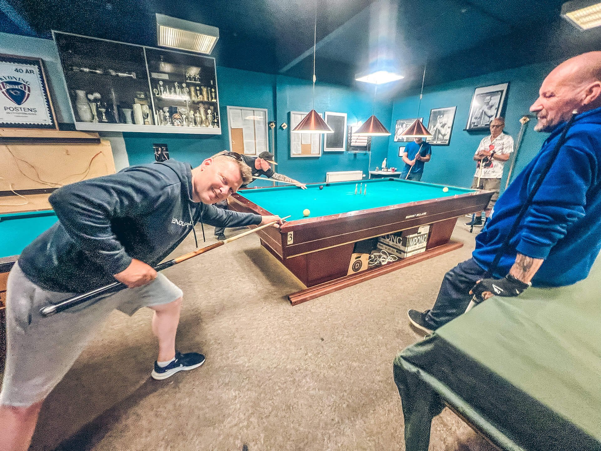 Mænd spiller billard i et klubhus med blå vægge og billeder på væggen.
