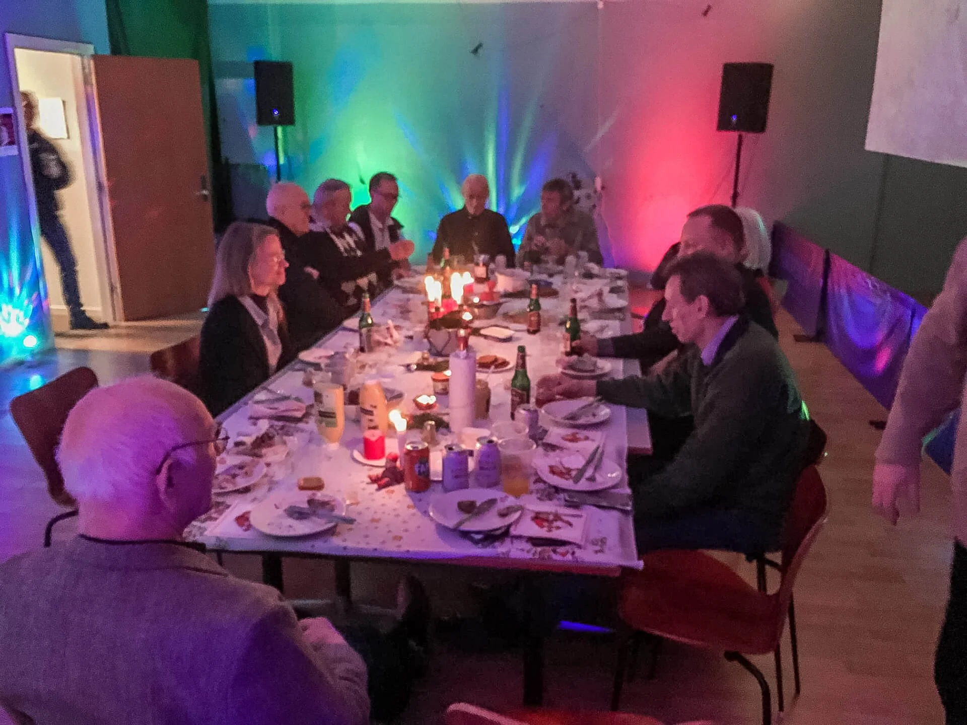 Mennesker sidder omkring et langbord dækket med mad og drikke ved en fest, med farvet belysning og stearinlys.