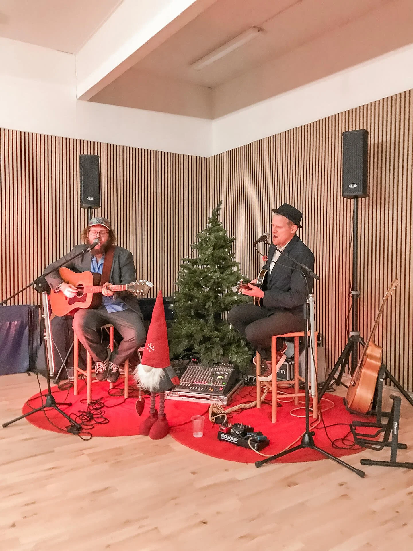 To mænd spiller musik foran en pyntet juletræ med nisser som dekoration. Den ene spiller guitar og synger, den anden spiller også guitar.