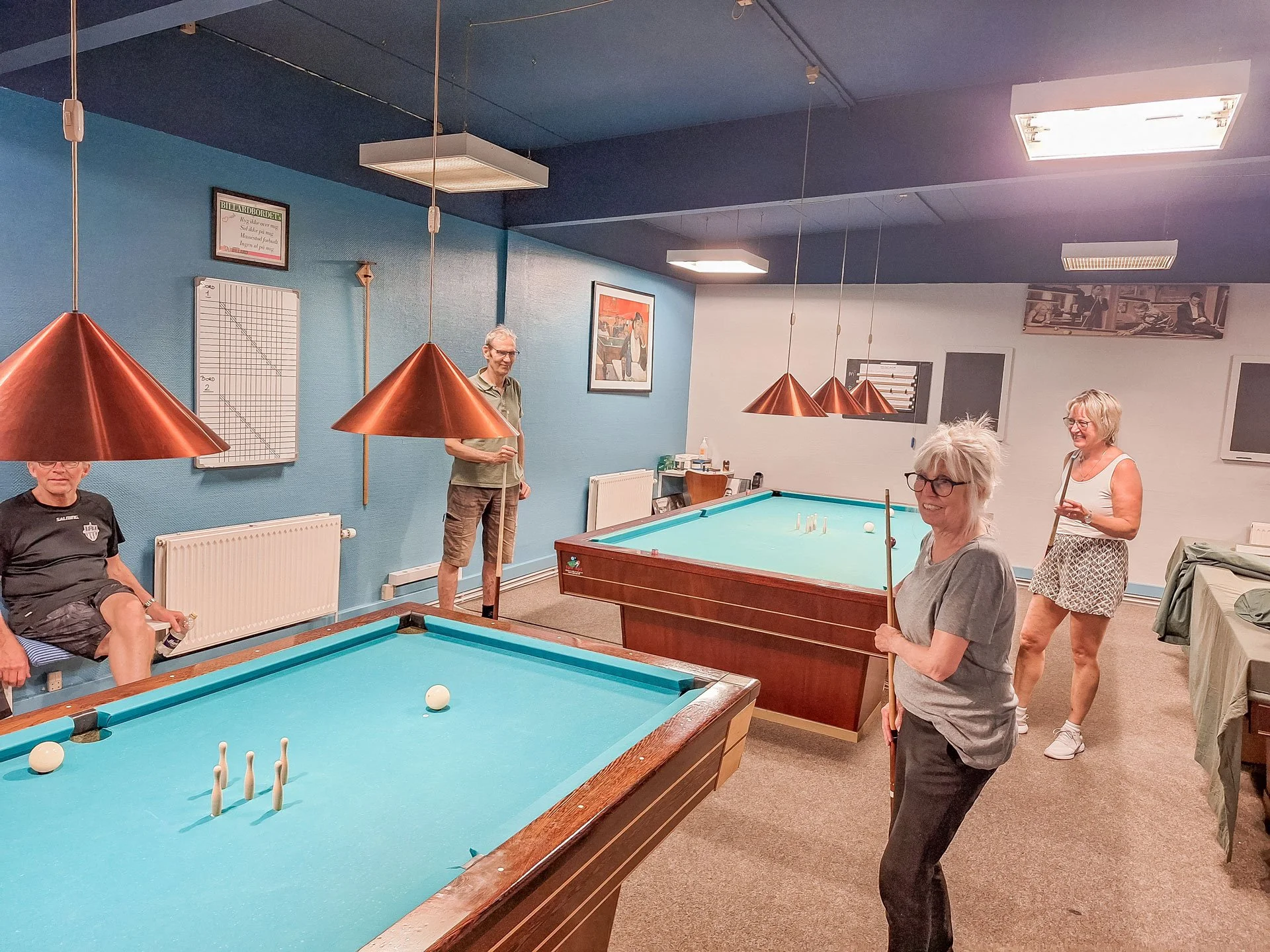 Fem voksne personer spiller billard i et rum med blå og hvid væg, kobberlamper og billeder på væggene.