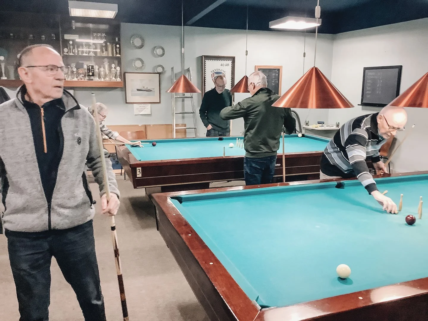 En gruppe ældre mænd spiller billiard i en hyggelig stue med poolborde, kobberlamper og ballonskabe på væggen.