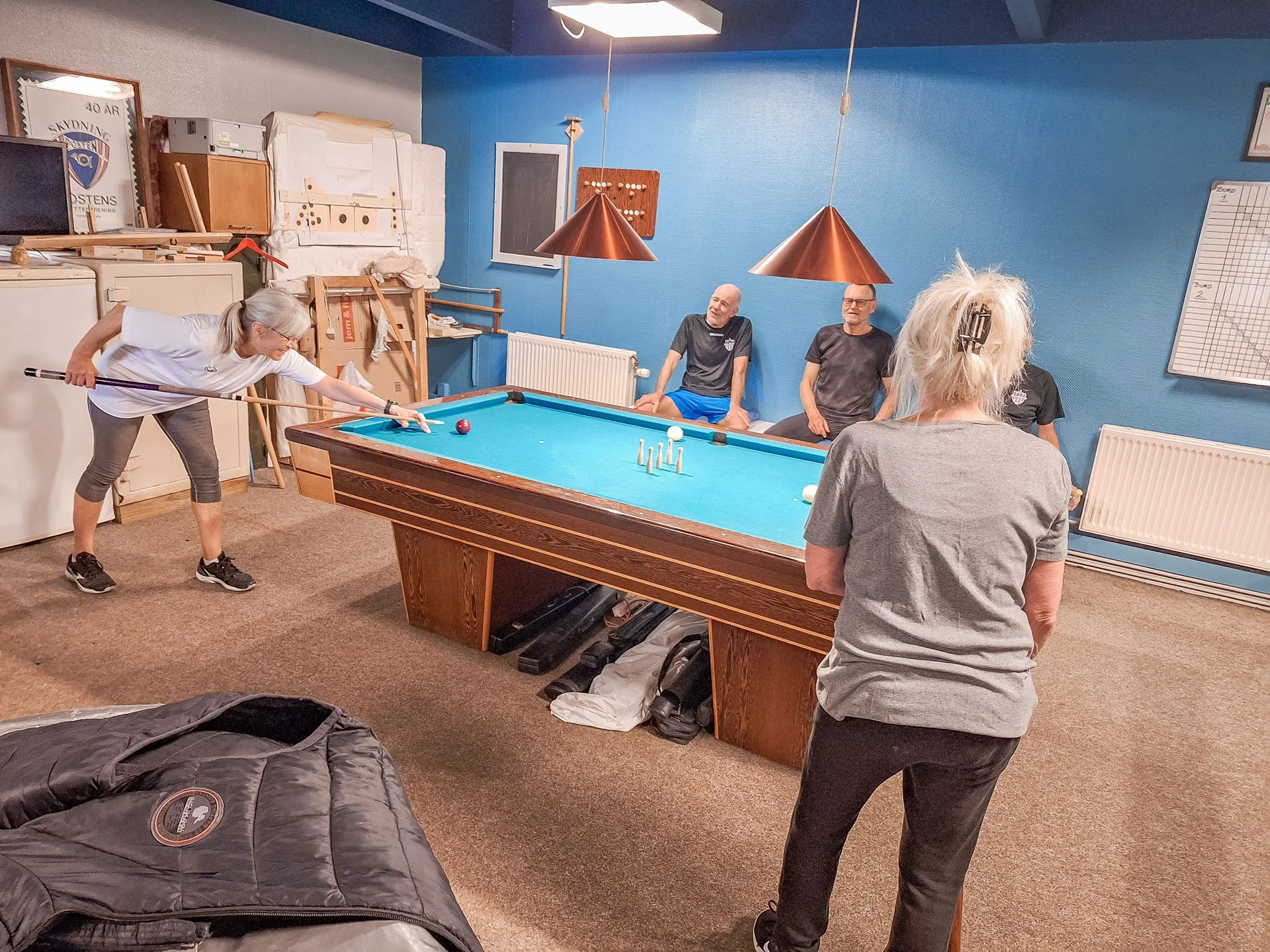 En gruppe mennesker spiller billard i et rum med blå væg. En kvinde skøder poolbolden, mens tre personer sidder og ser på.