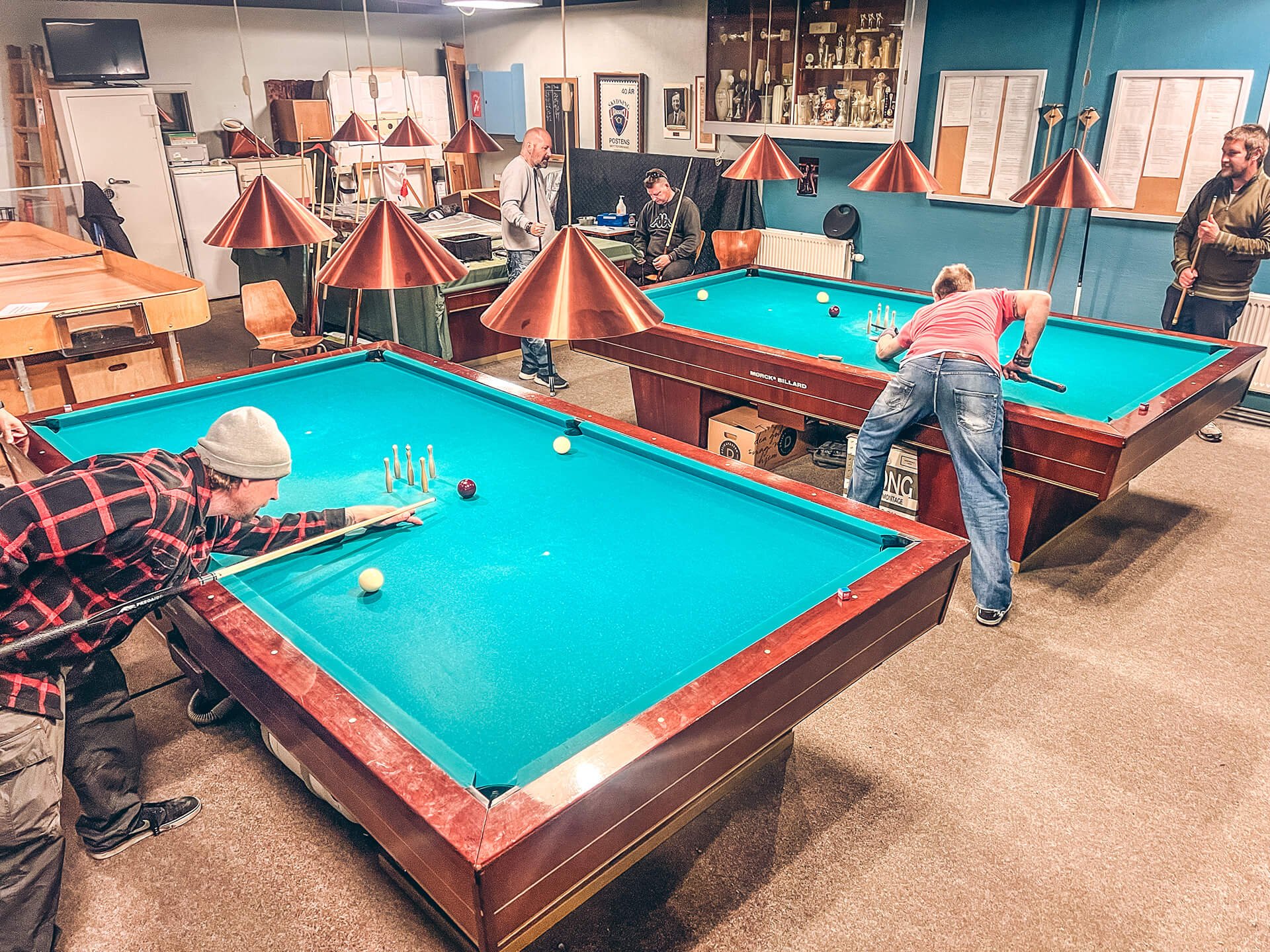 Mennesker spiller billard i en indendørs spillehal, med træborde og kobberlamper, samt hylder med trofæer og billeder på væggene.