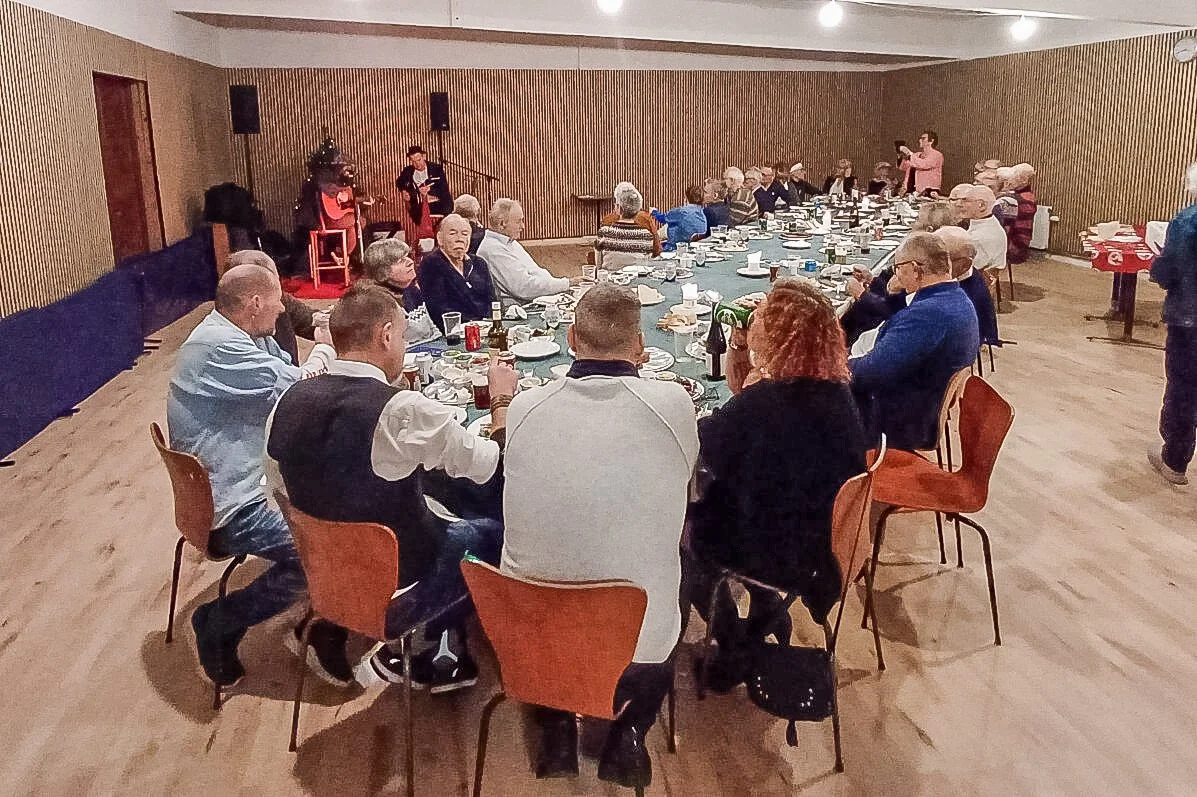 En gruppe mennesker sidder omkring et langt bord i et festlokale, mens nogle børn optræder på en scene i baggrunden.