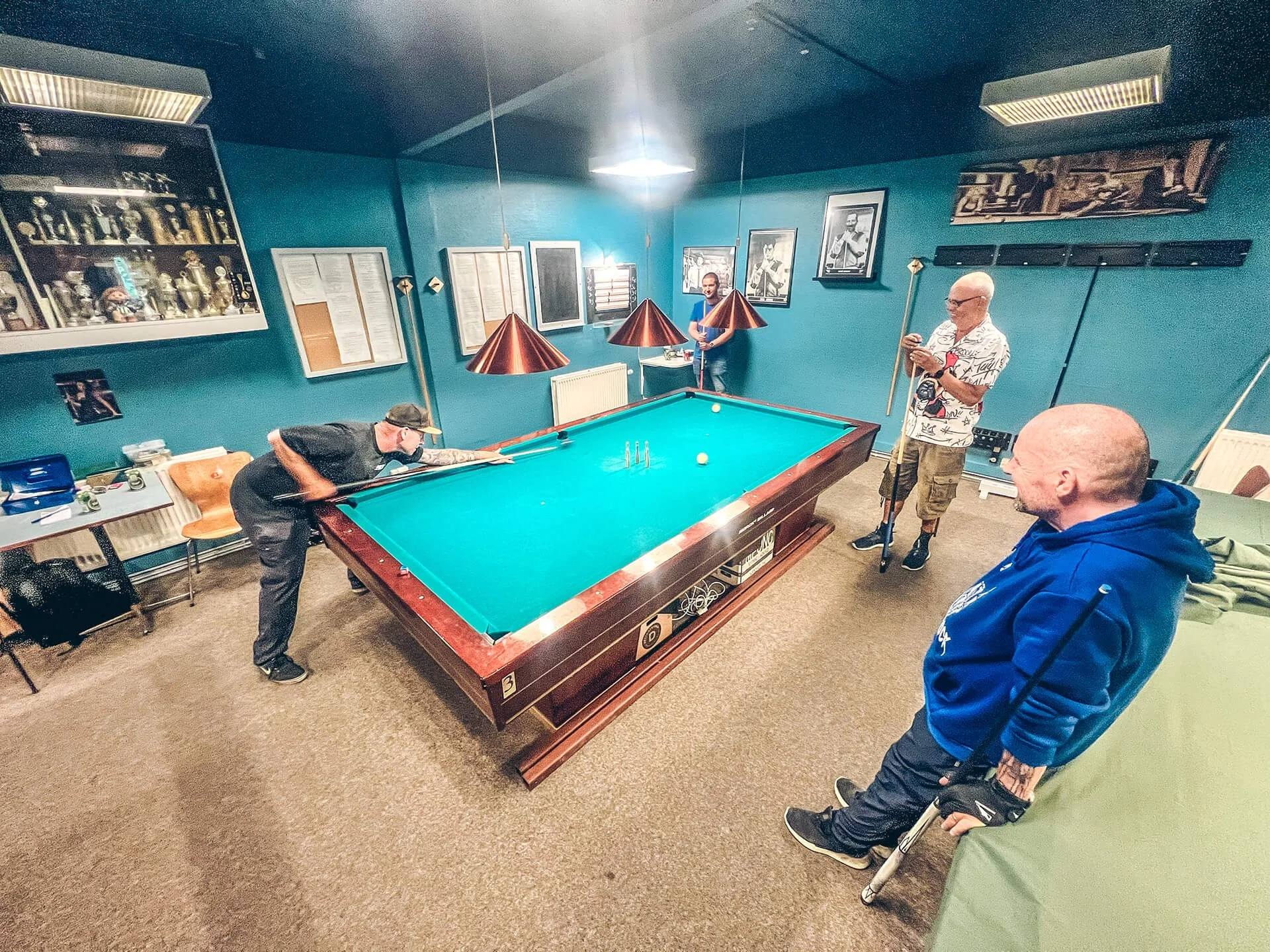 Tre personer spiller billard i et rum med blå vægge og fotografier på væggene.