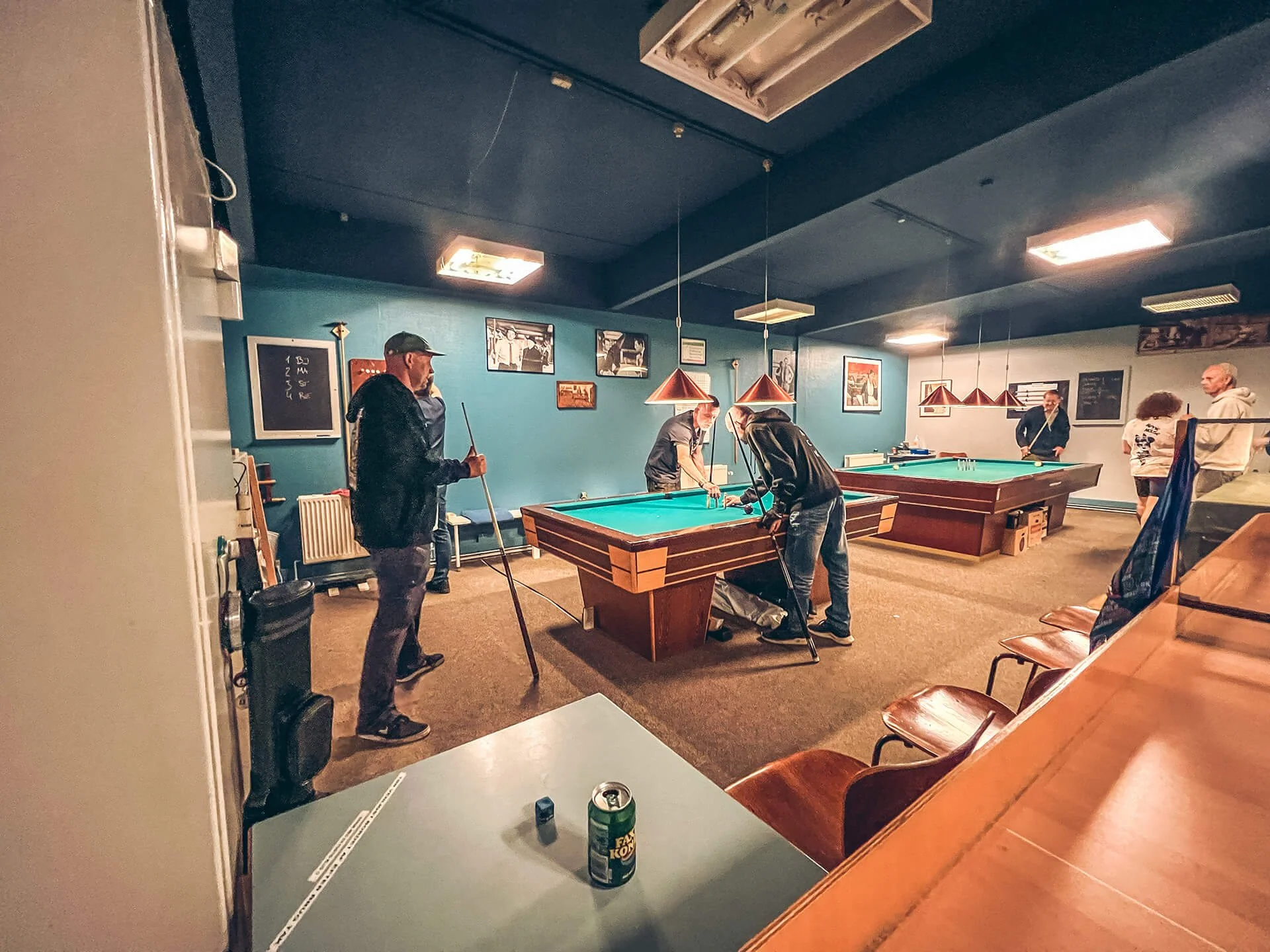 Mennesker spiller billard i et billardrum med blå vægge og billeder på væggen, bordlamper hænger over billardborde.