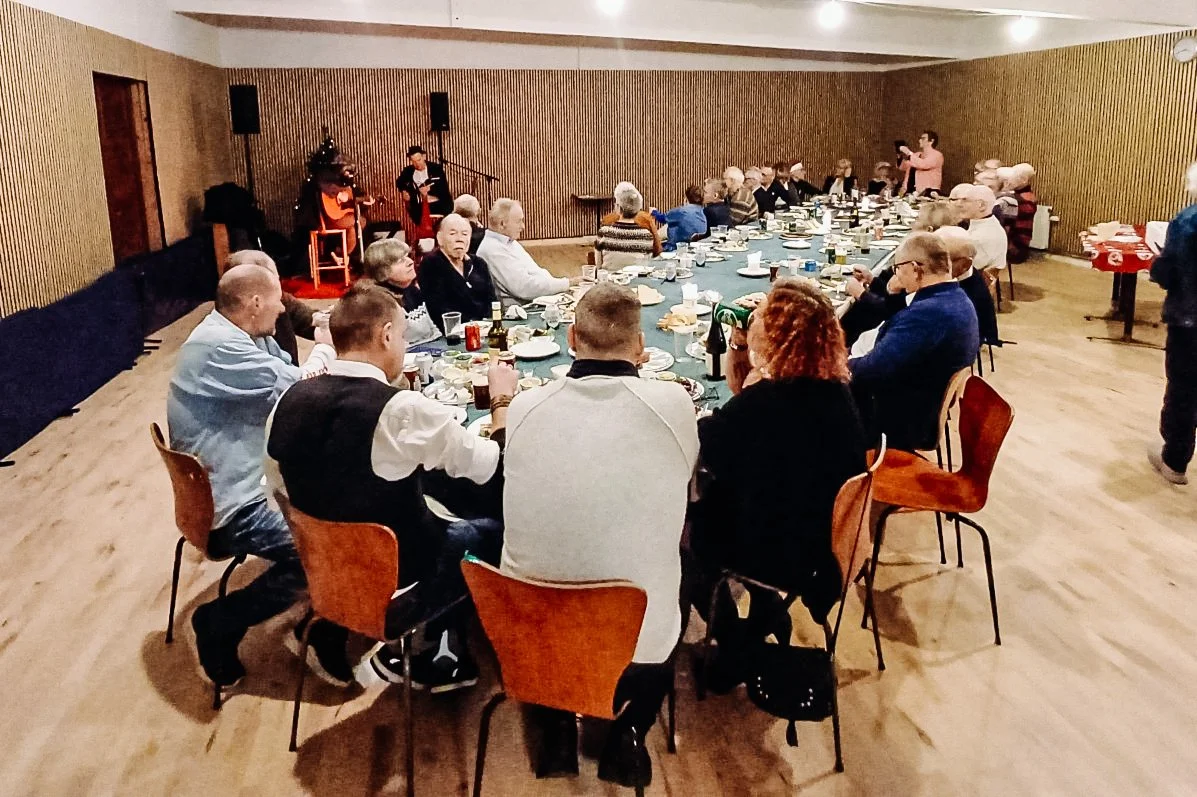 Folk sidder ved langbord med mad og drikke, mens der optrædes musik på scenen i baggrunden i en sportsklub.