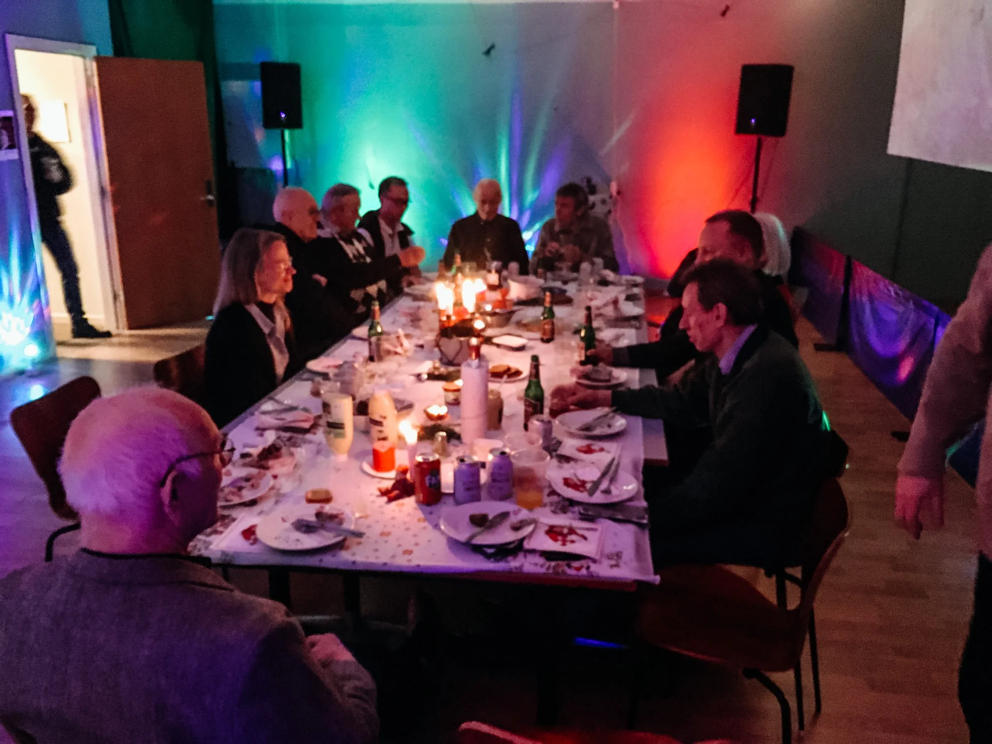 Personer sidder rundt ved et langbord, der er dækket med tallerkener, glas, bestik, lys og mad, i et festlokale med farvet lys.