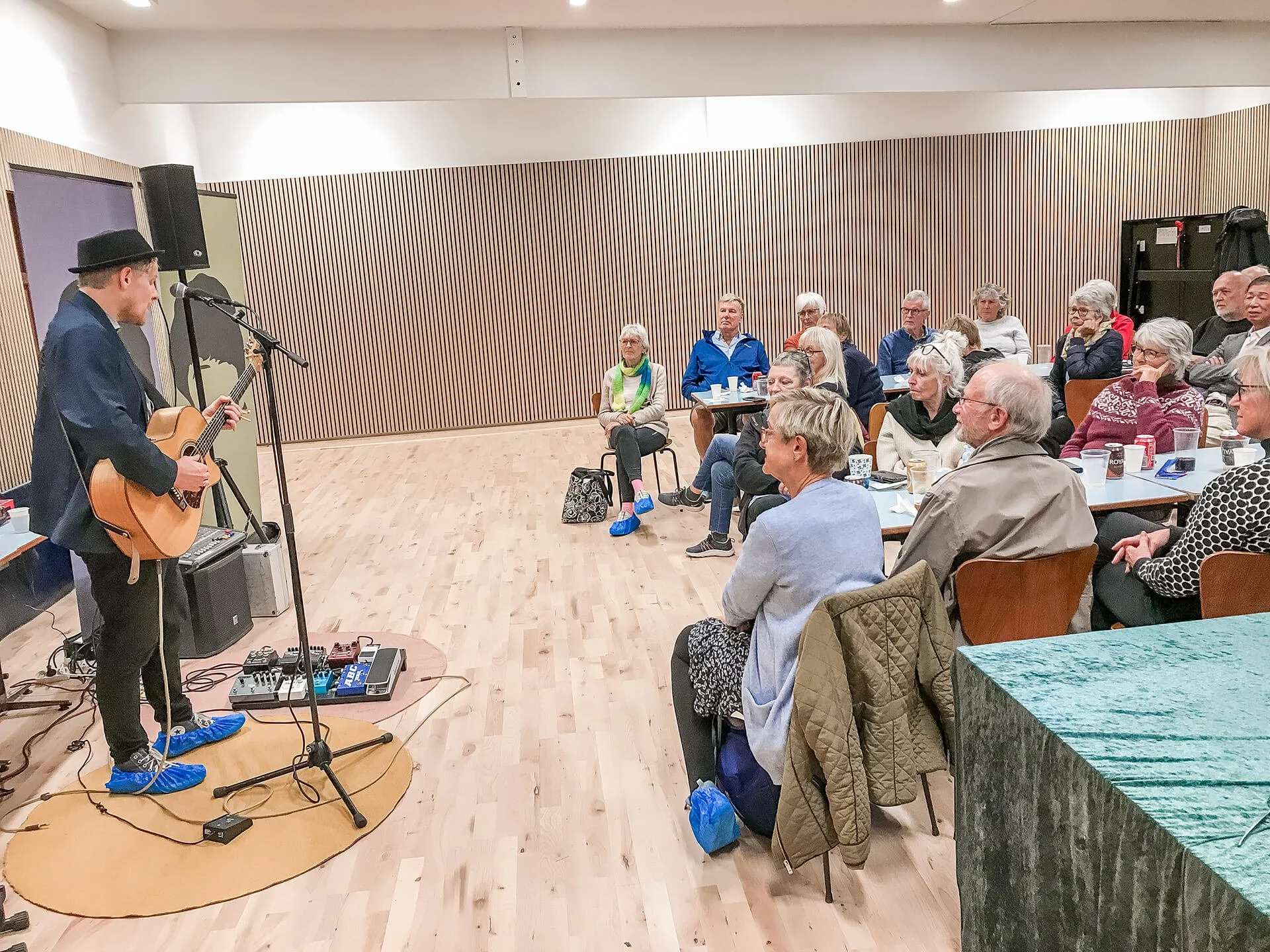 En mand spiller guitar for et publikum, der sidder ved borde i et foreningslokale.