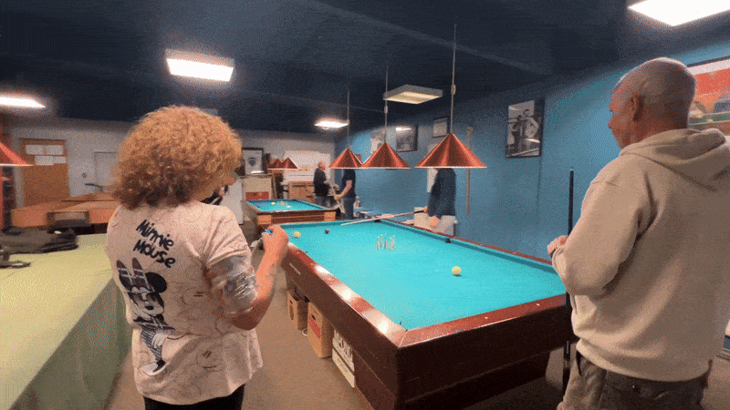 To personer spiller billard i en hyggelig lounge, med flere andre personer i baggrunden.
