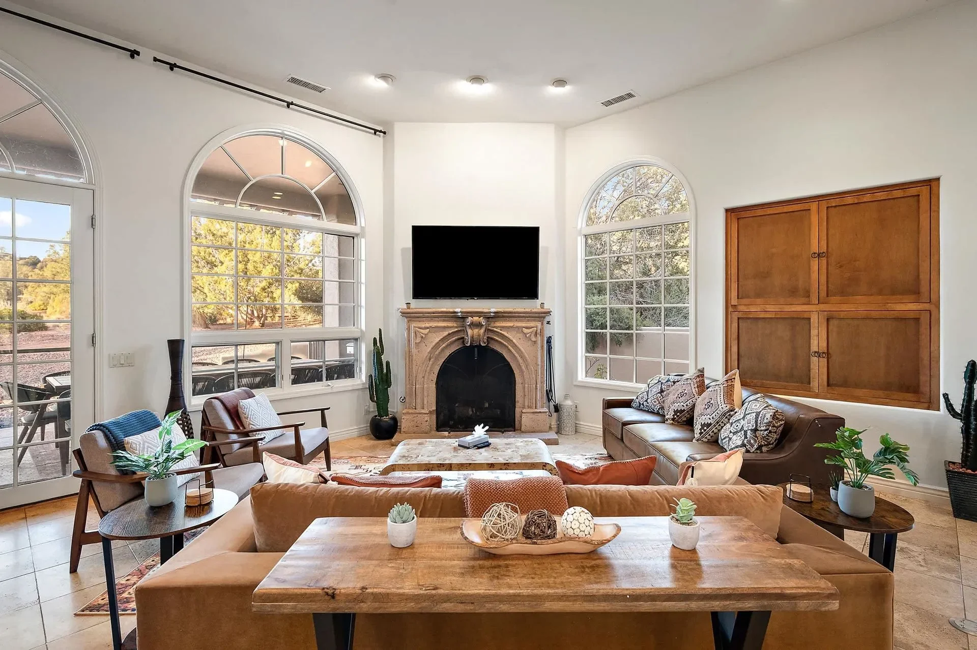 Living room with large windows, a fireplace, a mounted TV, and various seating including a sofa and armchairs, decorated with plants and cushions.