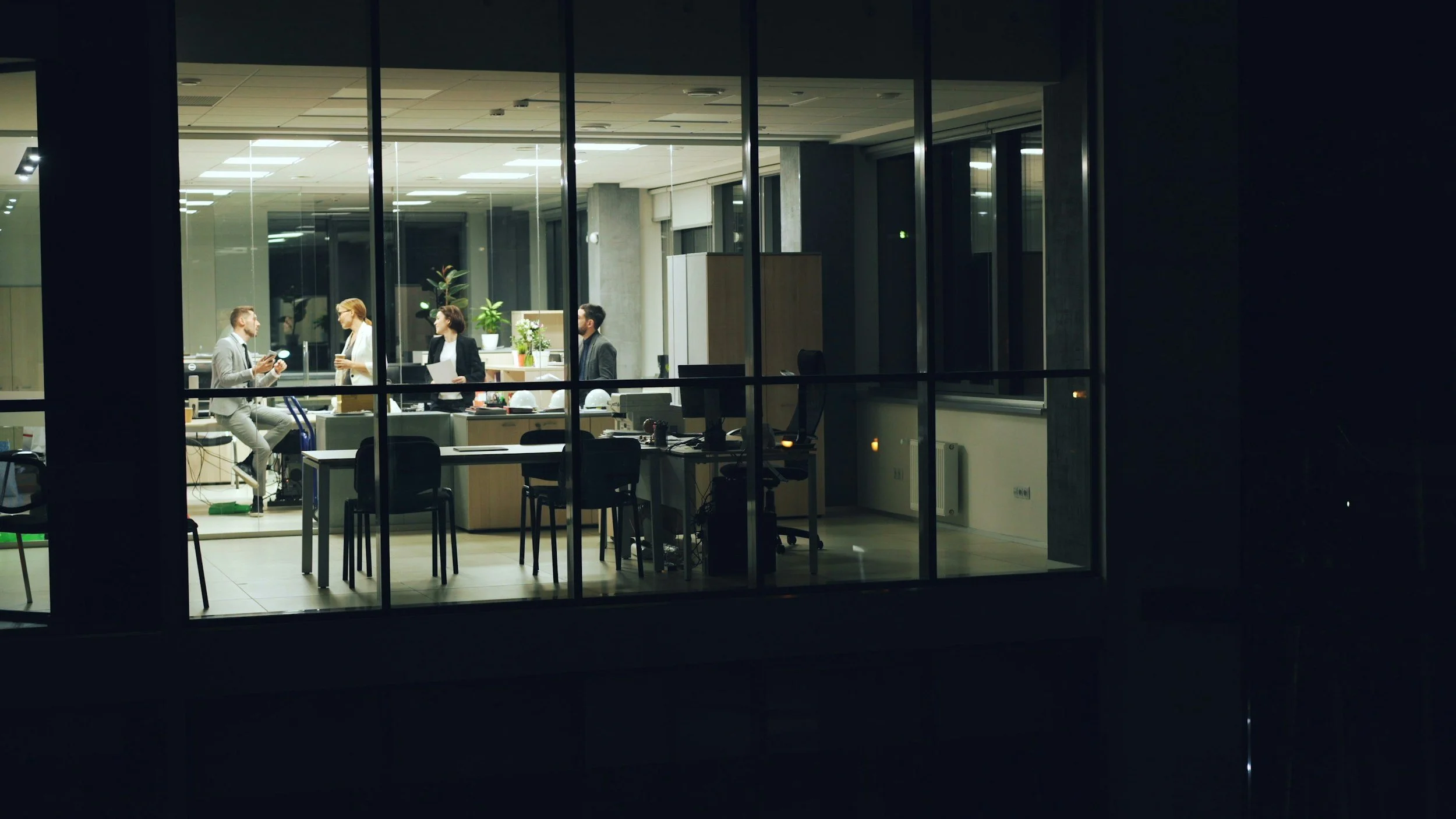 View through office window showing five people conversing inside an office at night, with desks, chairs, and plants visible.