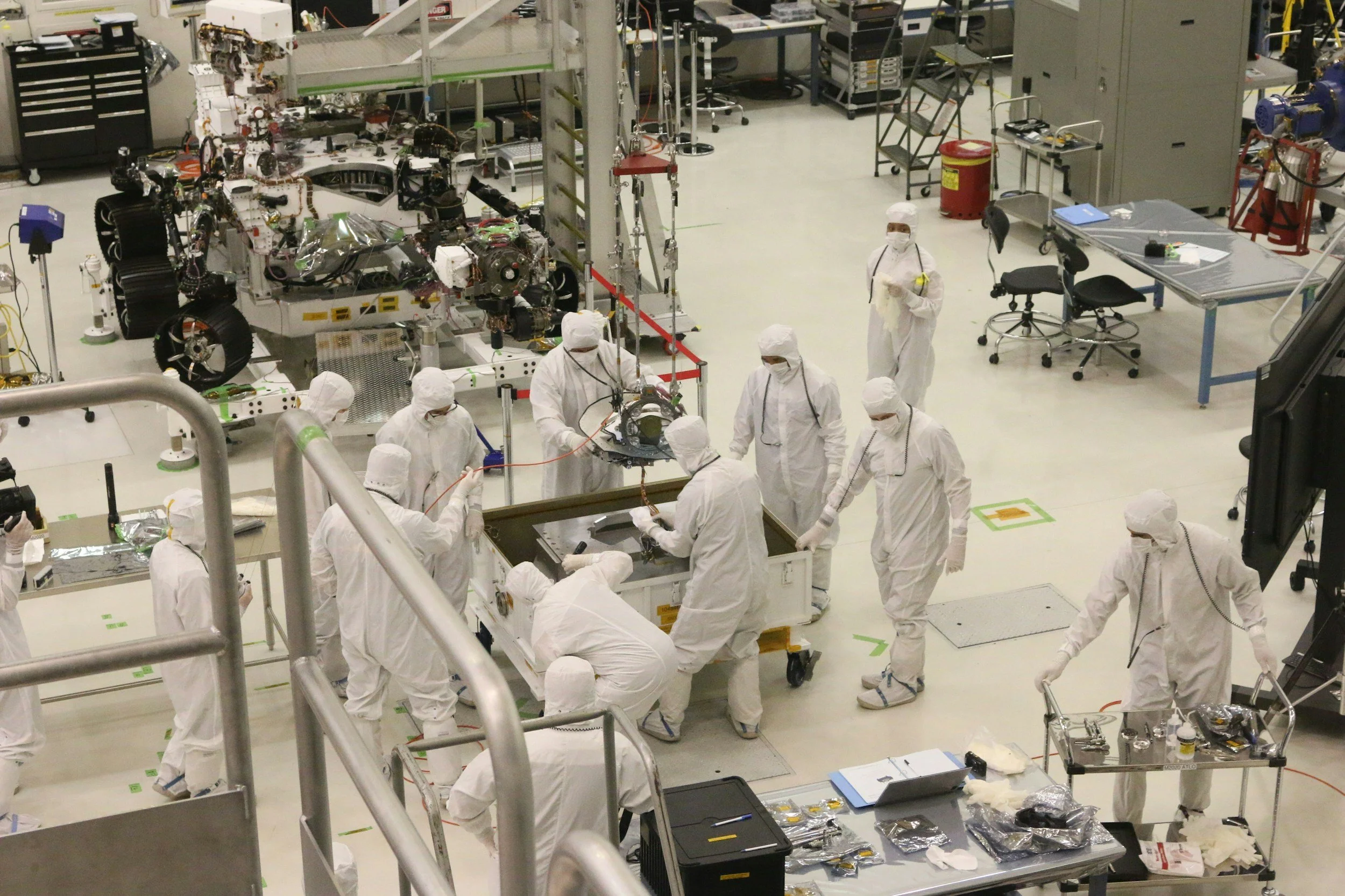 Scientists in white protective suits working in a cleanroom, assembling a spacecraft or satellite, surrounded by equipment, tools, and robotic arms.
