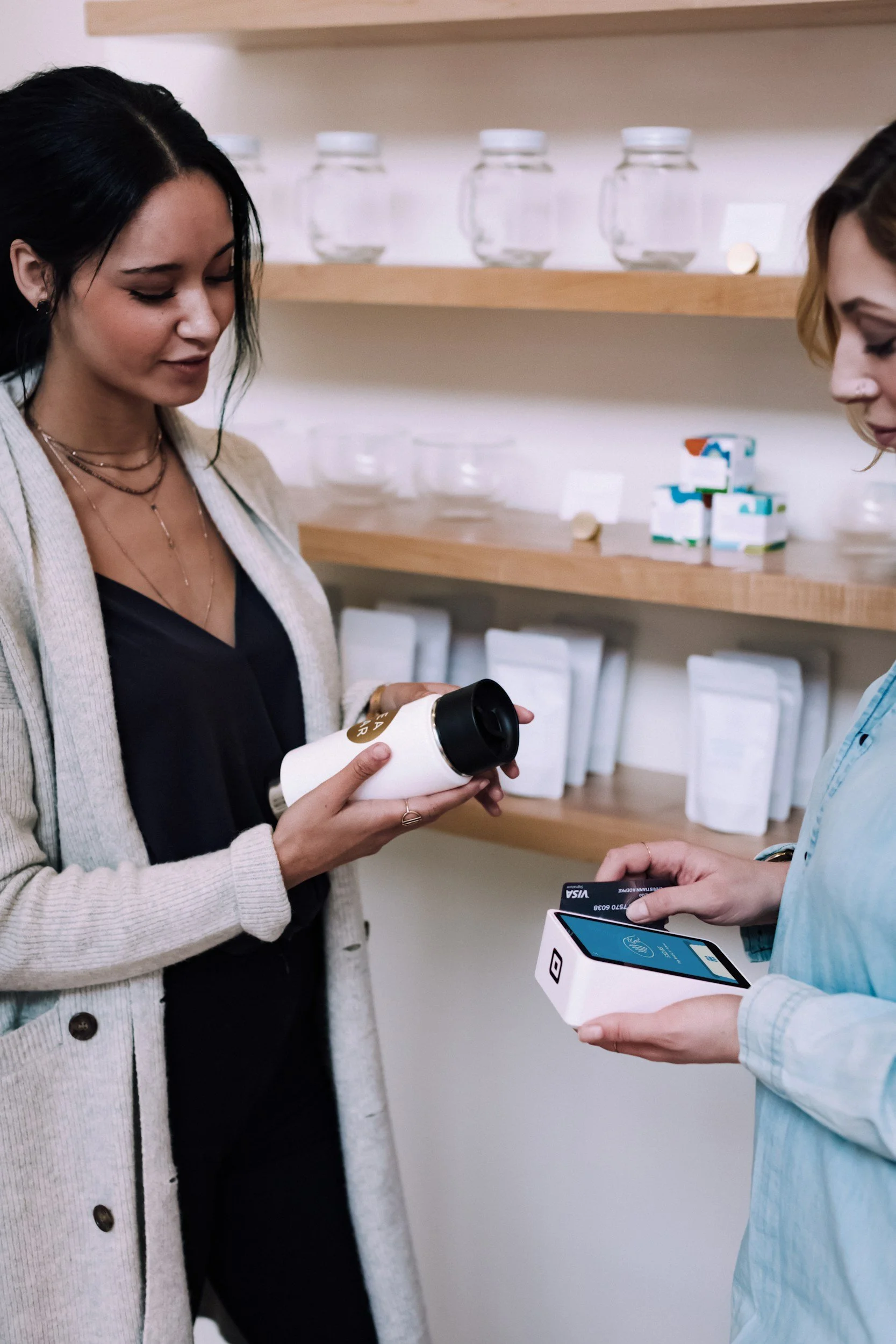 Two women shopping in a store; one woman is holding a white tumbler and looking at it, while the other woman is holding a mobile payment device and a card.