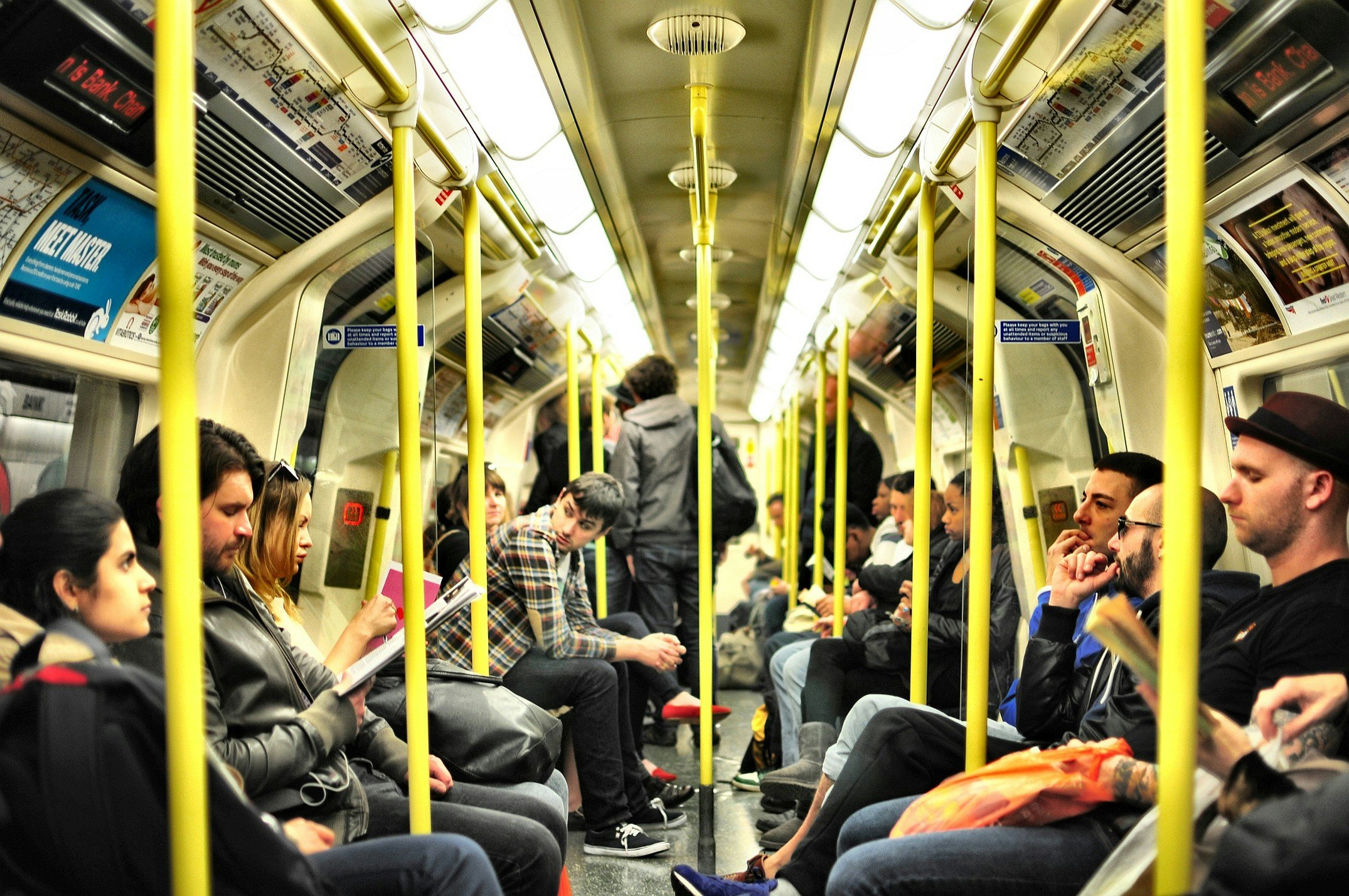 Inside a subway train with passengers seated and standing, yellow poles for support, advertisements above the windows, and fluorescent lighting.