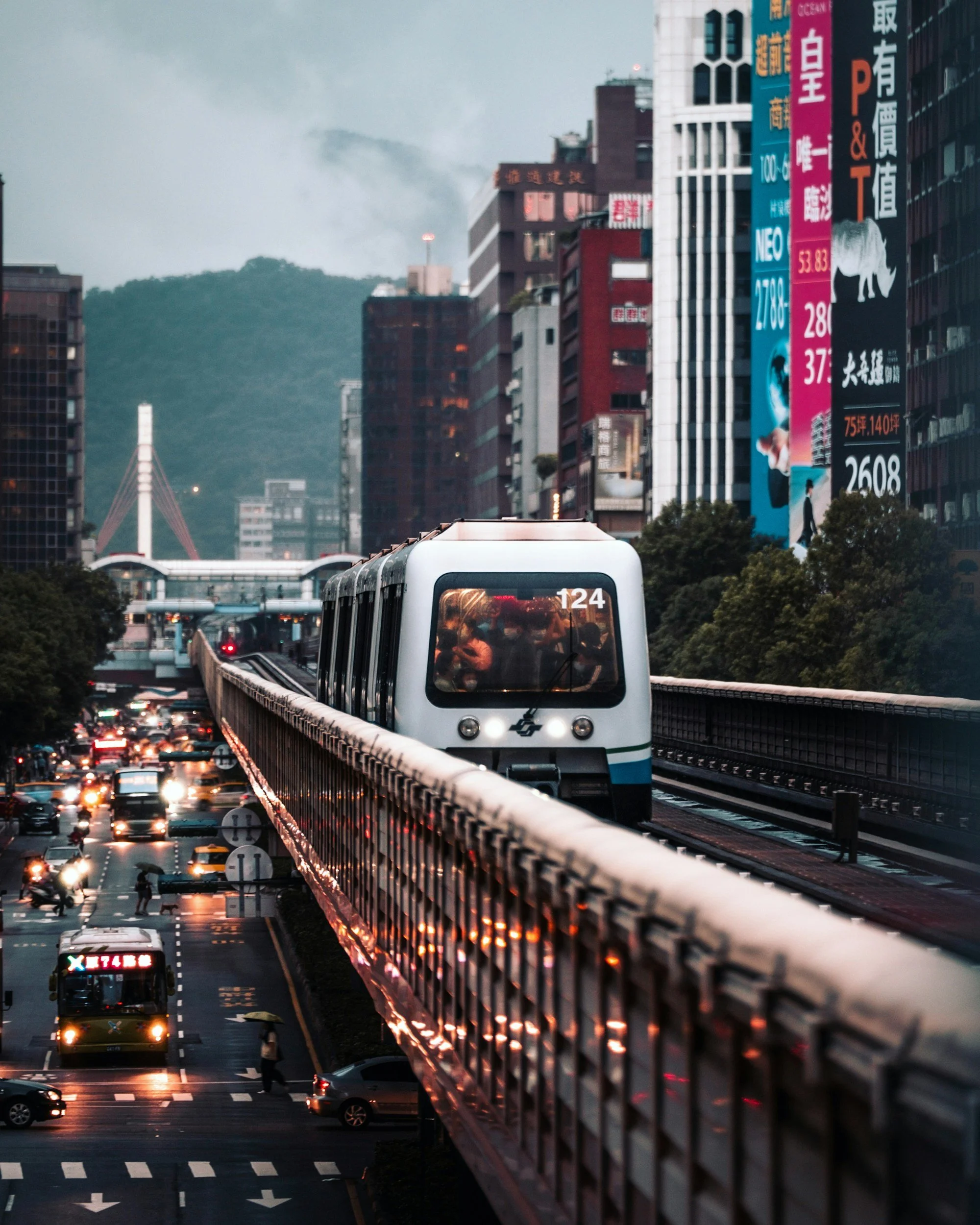 Urban scene with elevated train traveling above busy street with cars, buses, and pedestrians, surrounded by high-rise buildings with colorful advertisements in the background.