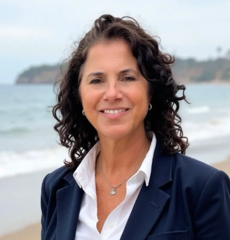 A woman with curly dark hair smiling at the camera on a beach with ocean waves and a distant landmass in the background.