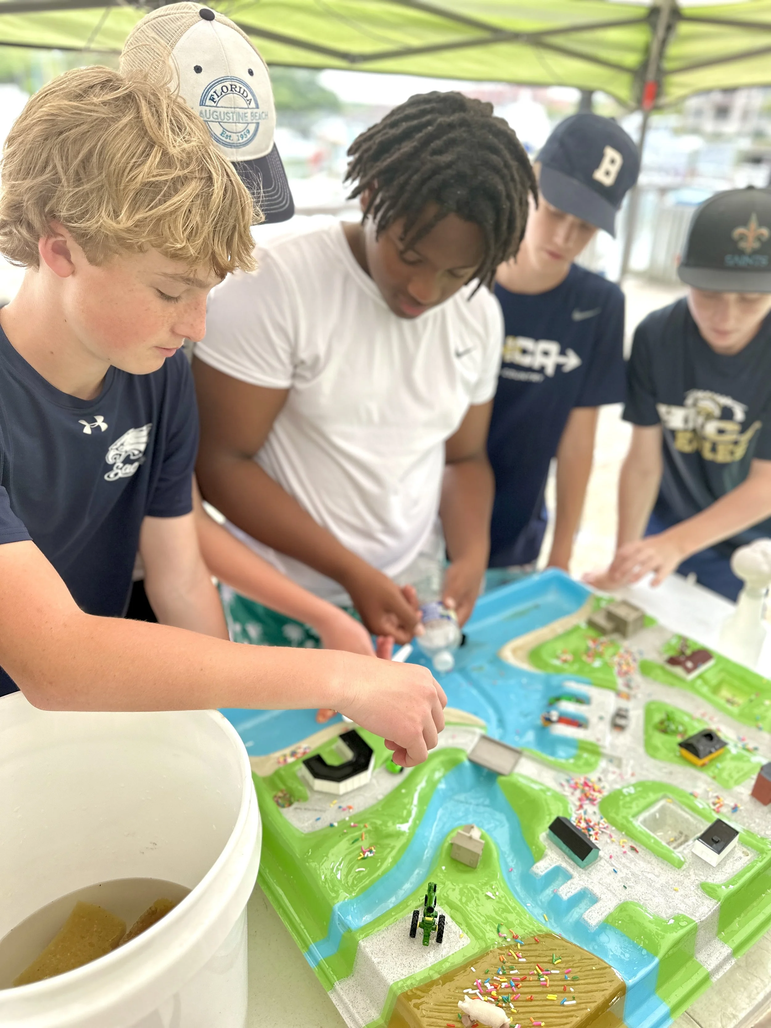 Group of young people playing with a colorful tabletop game under a green canopy. Various game pieces, figures, and decorations on game boards are visible.