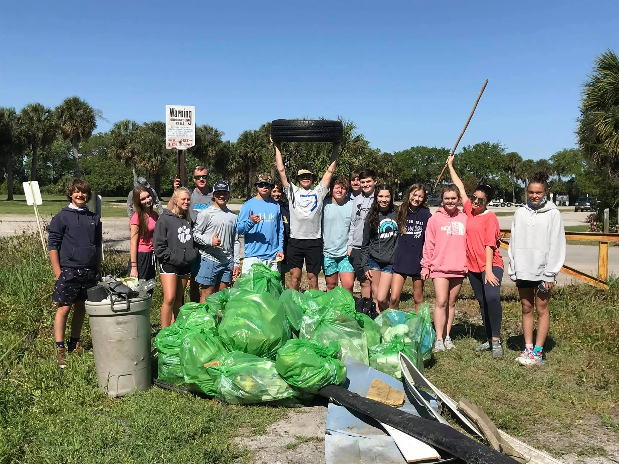 A group of young people gathering outdoors for a cleanup activity. They are standing behind a pile of green trash bags filled with collected litter. The background shows a bright, sunny day with blue skies, palm trees, and some signage. The group appears to be smiling and enjoying the event.