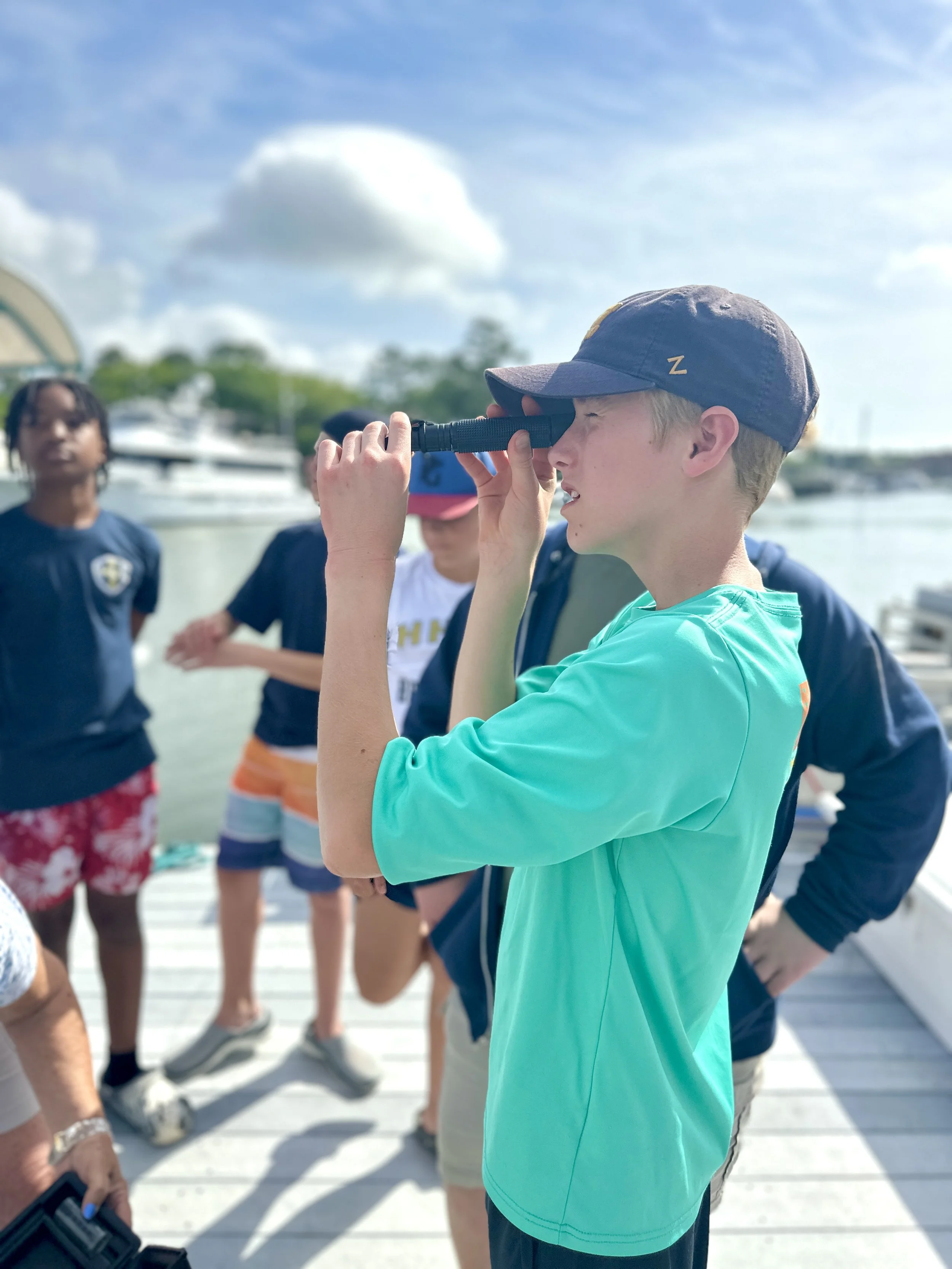 Young boy with blond hair and a dark cap looking through a telescope at a boat dock, with other children and boats in the background on a sunny day.