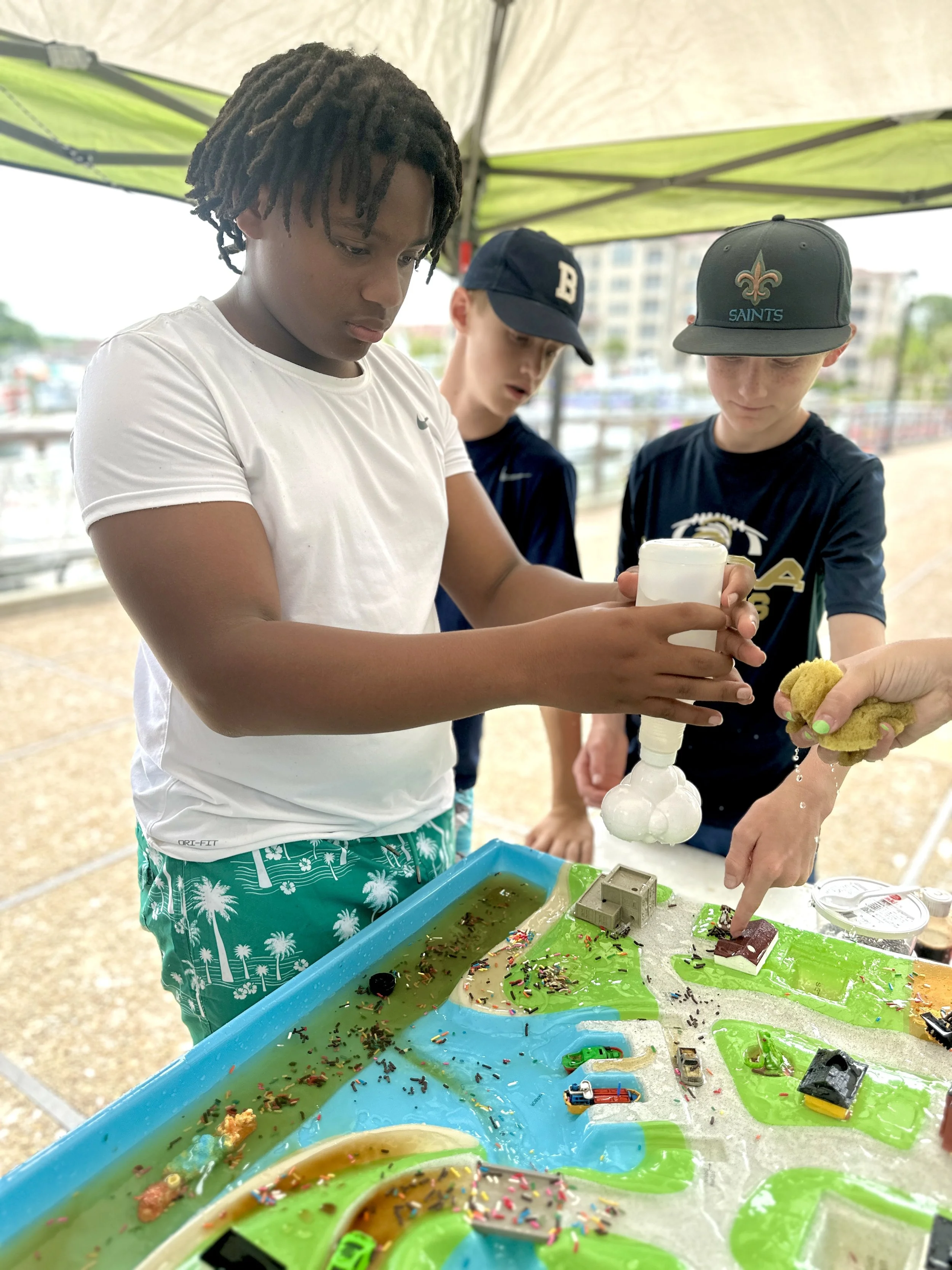 Three boys at a game table with green, blue, and yellow compartments filled with water and toys, some toy cars and chocolate, under a canopy, engaged in a game involving squeezing a white object and a sponge.