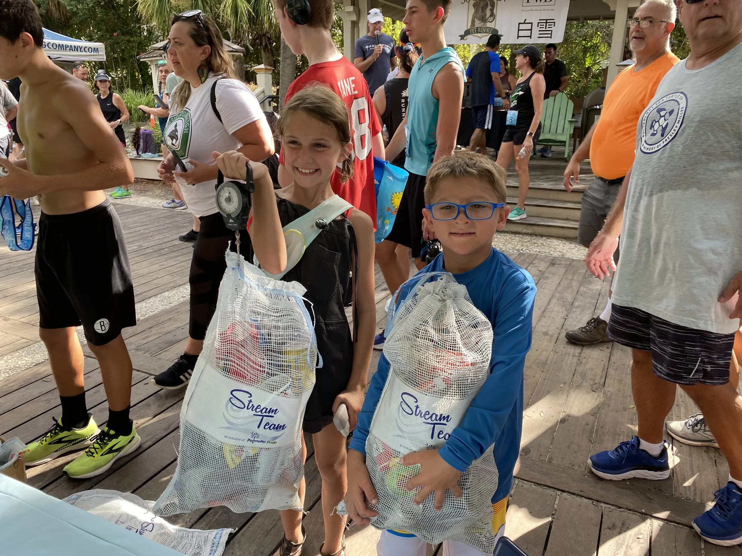 People participating in a marathon event, some are holding mesh bags, with a young girl and boy smiling in the foreground, and a crowd of runners and spectators behind them.