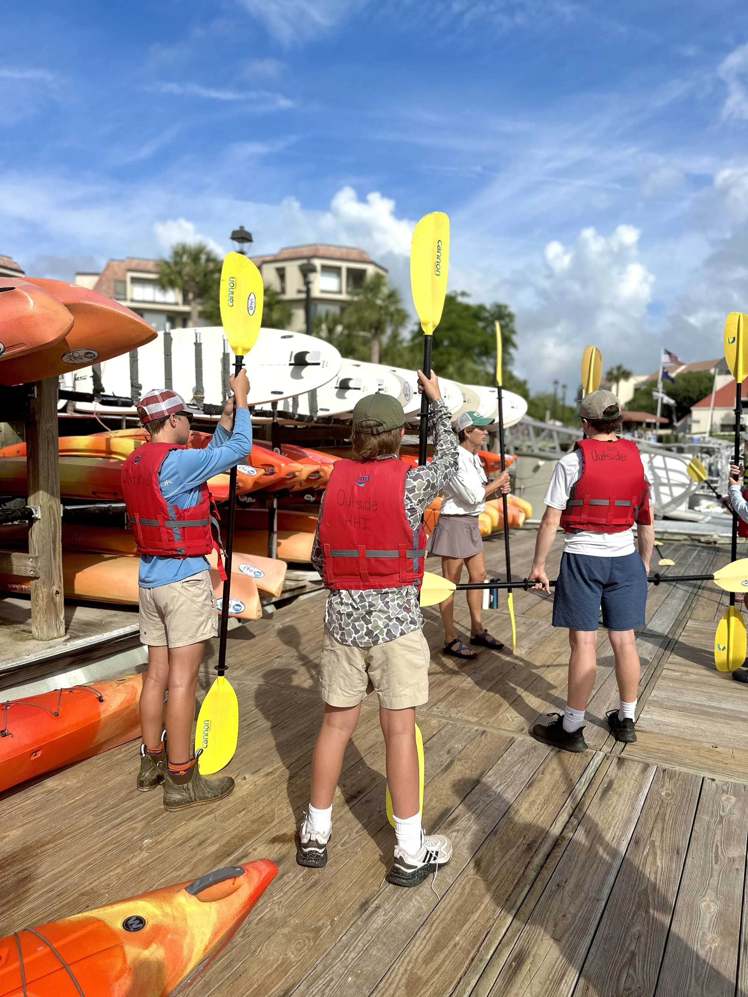 People wearing life jackets on a wooden dock, preparing for kayaking, with kayaks and paddleboards stored behind them, and blue skies with some clouds overhead.