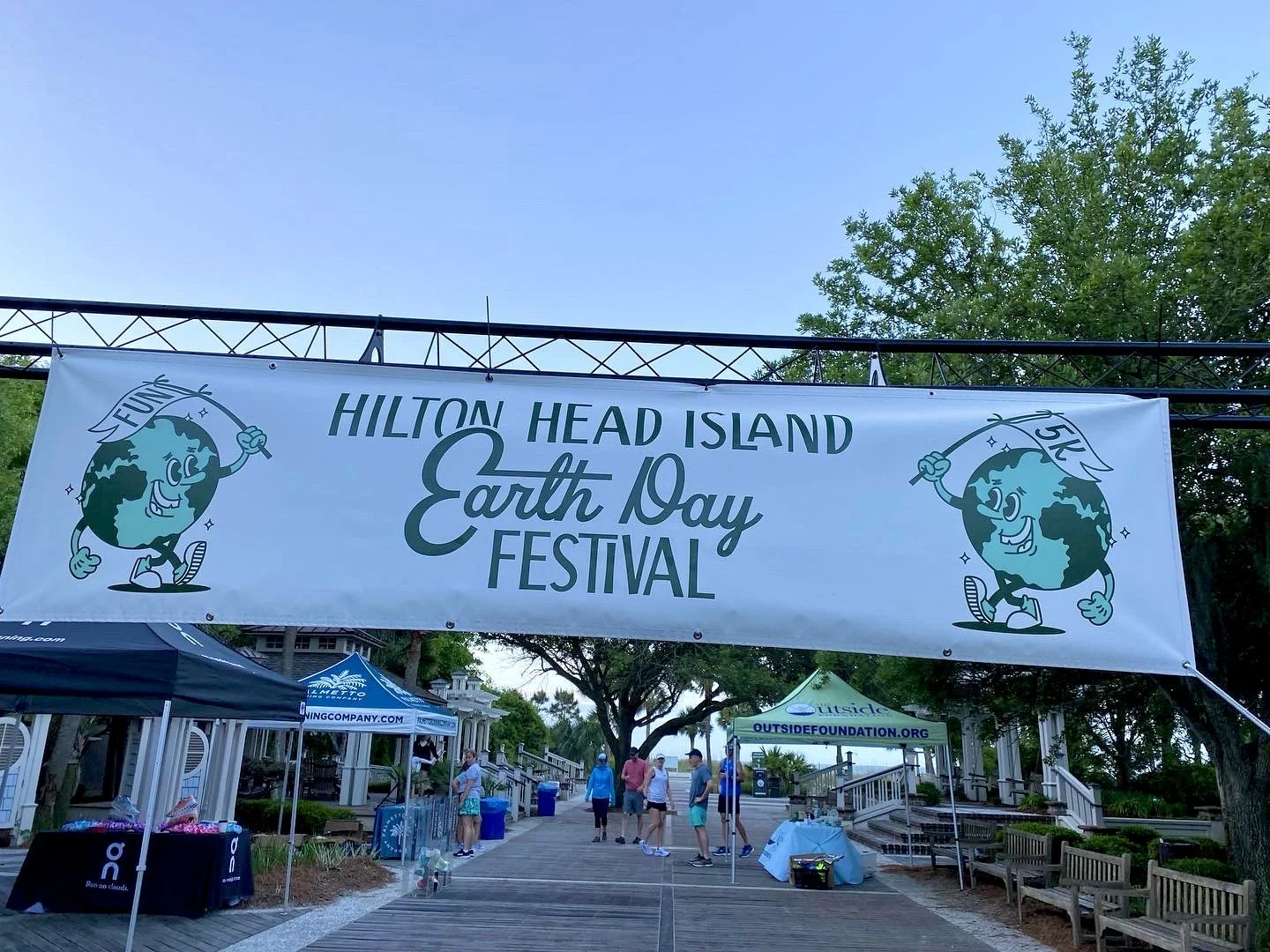 Banner for Hilton Head Island Earth Day Festival with cartoon Earth holding a flag that says 5K, and the words Hilton Head Island Earth Day Festival, with tents and people walking underneath.