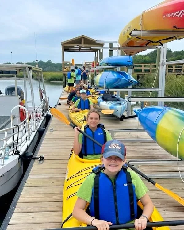 Kids preparing for kayaking on a wooden dock with kayaks and colorful canoes, alongside a boat in a natural water setting.