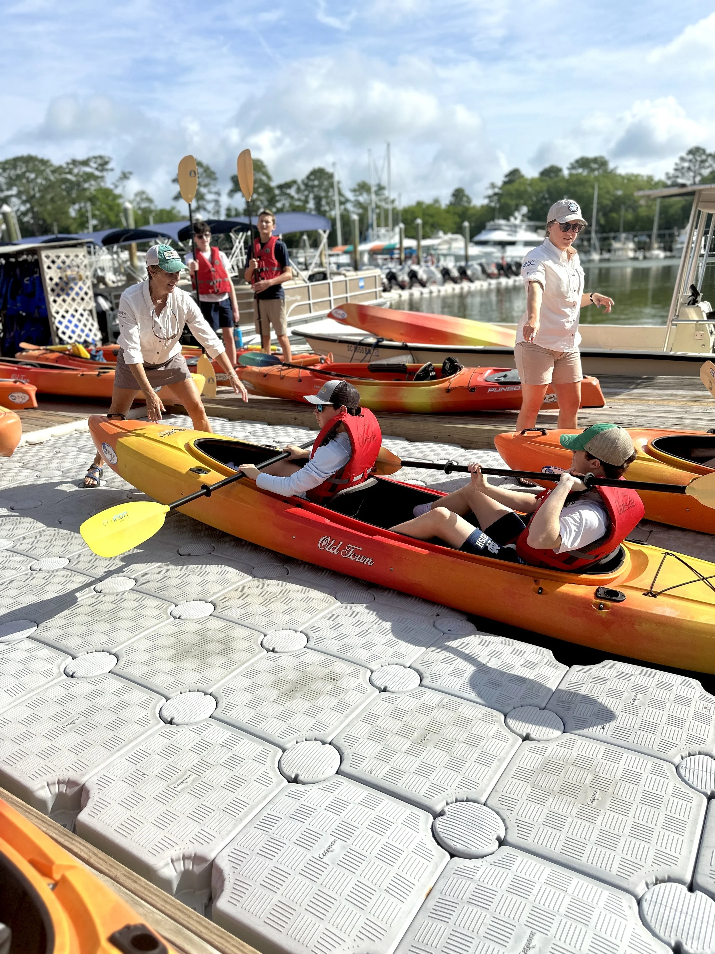 People preparing kayaks and paddleboards at a marina on a sunny day.