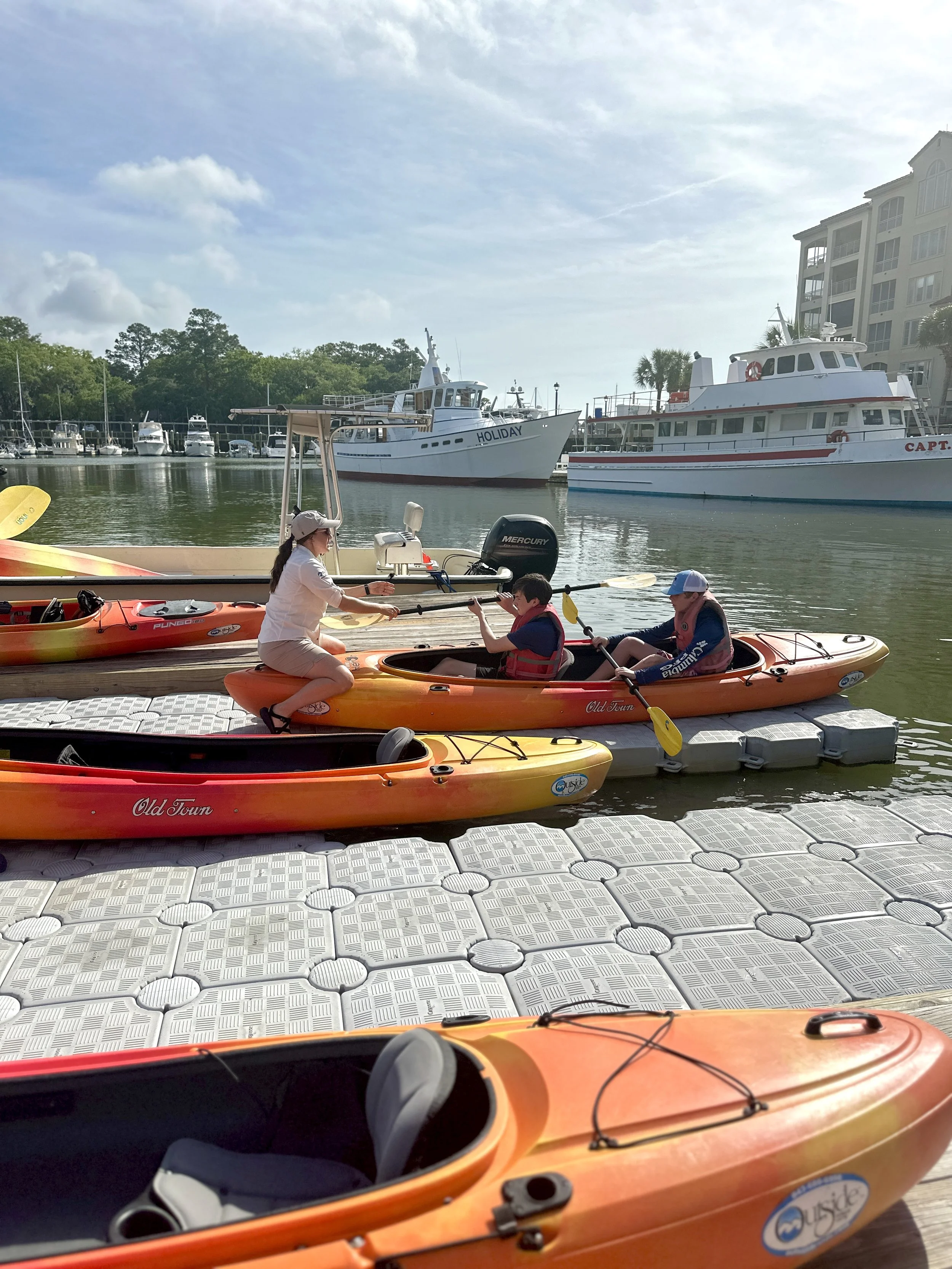 People preparing to kayak near a marina with boats in the background and a building on the right side.