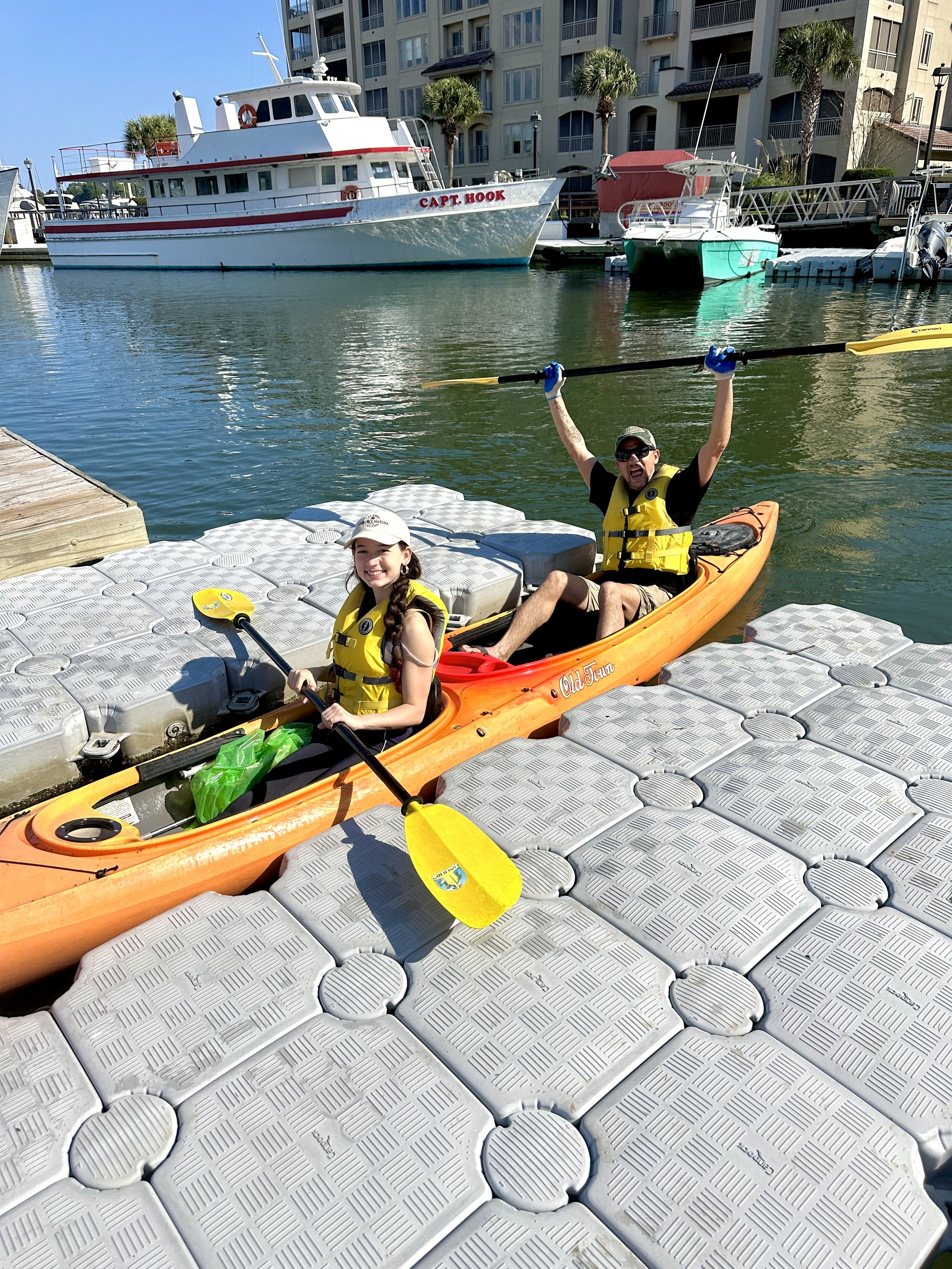 People in a kayak at a dock with a marina and boats in the background.