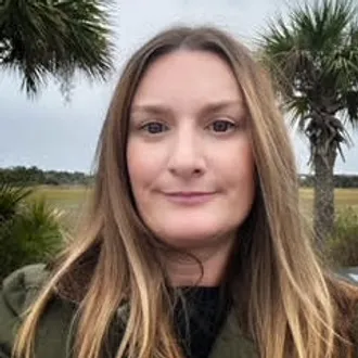 A woman with long, light brown hair stands outdoors with palm trees and a grassy field in the background.