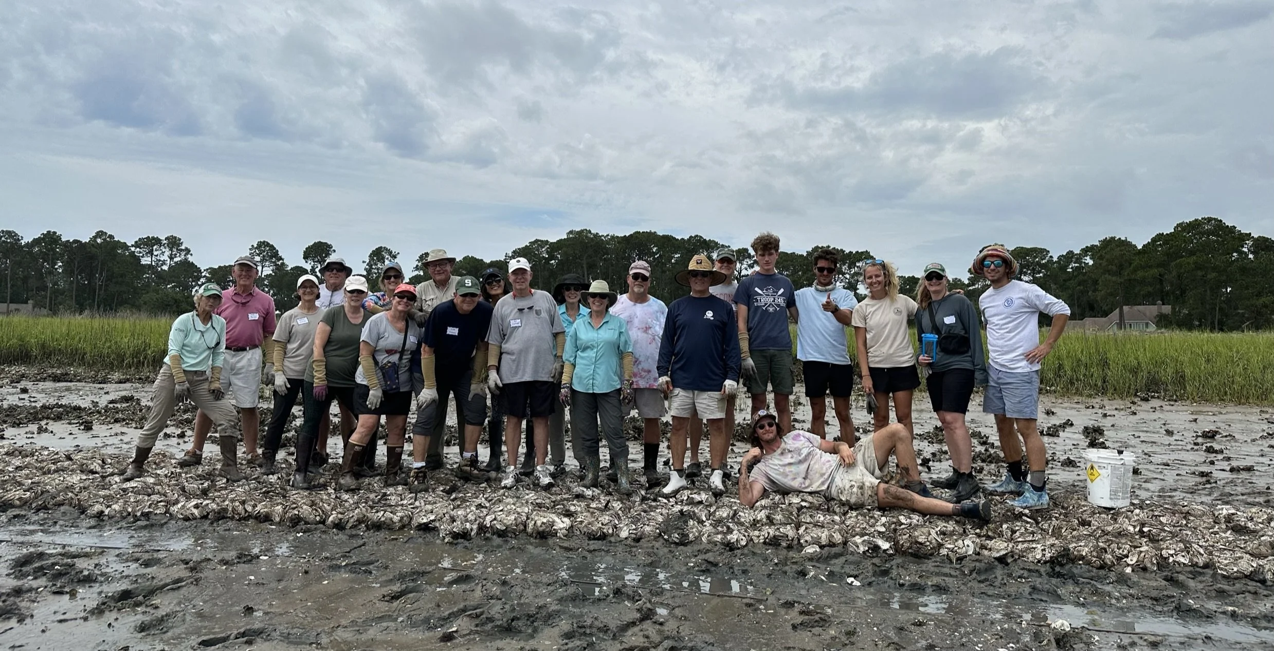 Group of people outdoors, some with gloves and boots, standing on a muddy and shell-covered shoreline with green marsh grass and trees in the background under cloudy skies.