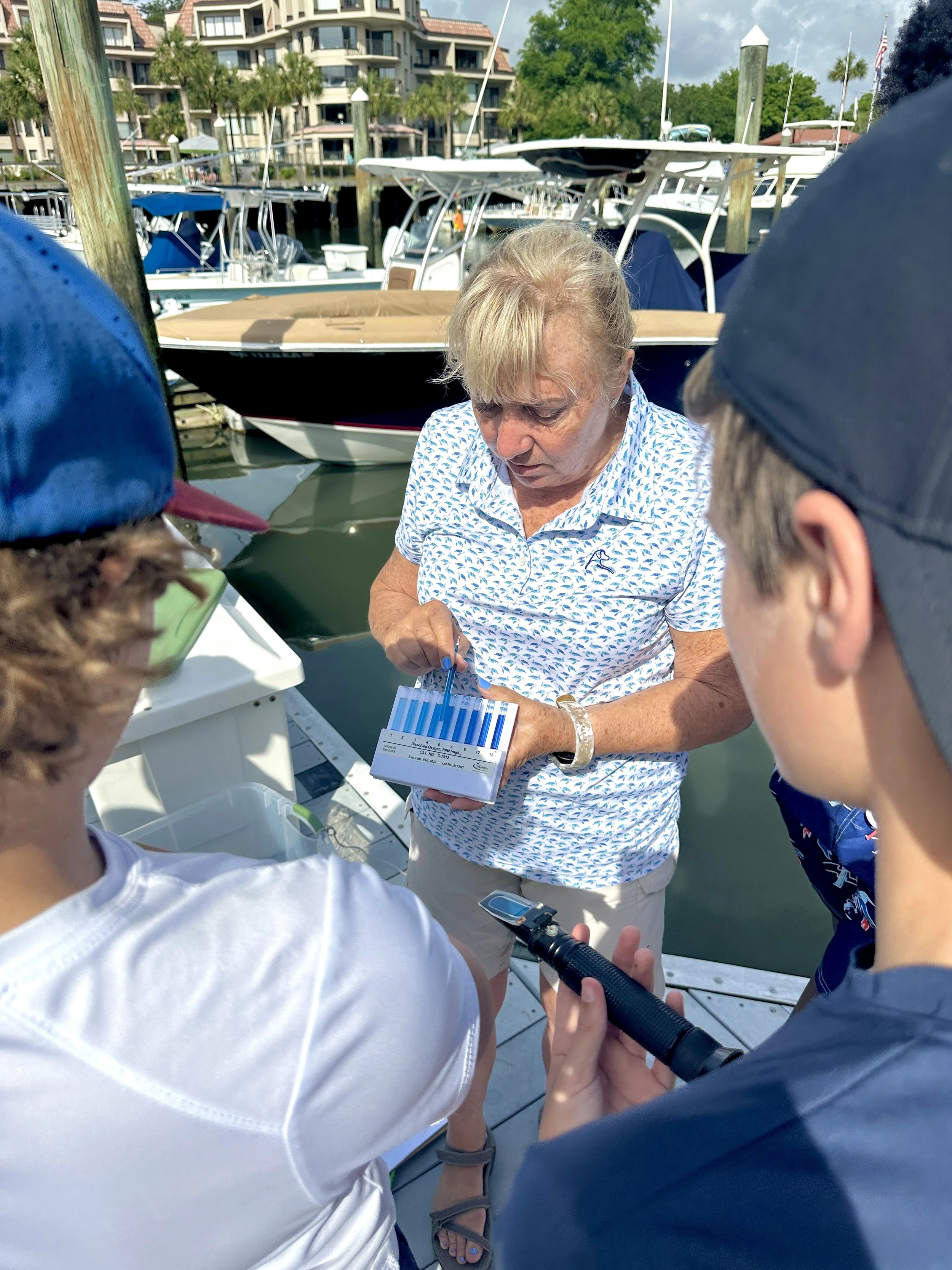 A woman at a marina with boats and waterfront buildings in the background, holding a box of blood test tubes and discussing with three children.