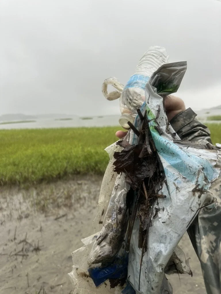 Hand holding a pile of trash, including a crumpled plastic bottle and other debris, in an outdoor muddy area with green fields and cloudy sky in the background.