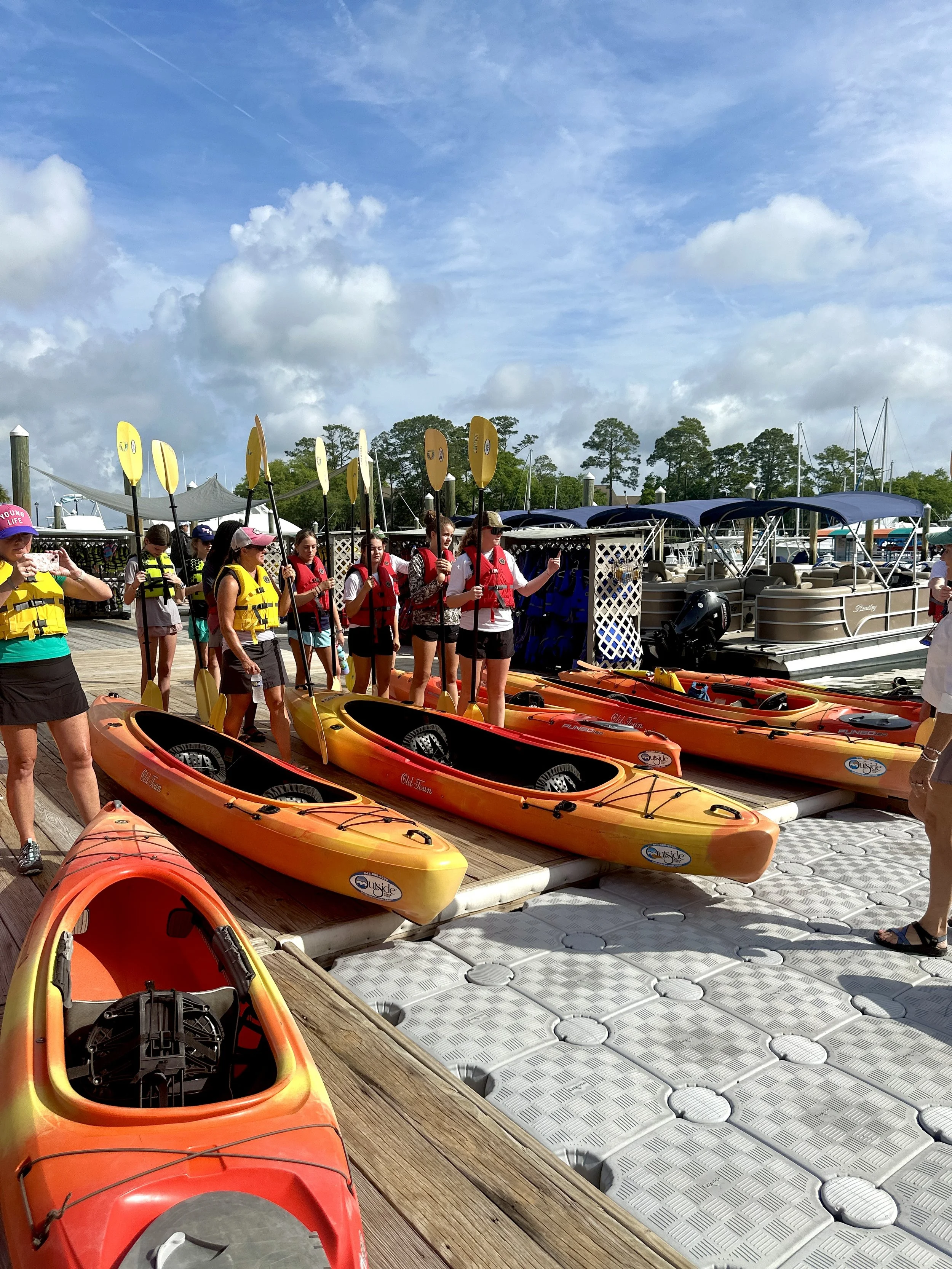 Group of people preparing to kayak at a marina dock, with yellow and orange kayaks and boats in the background under a partly cloudy sky.