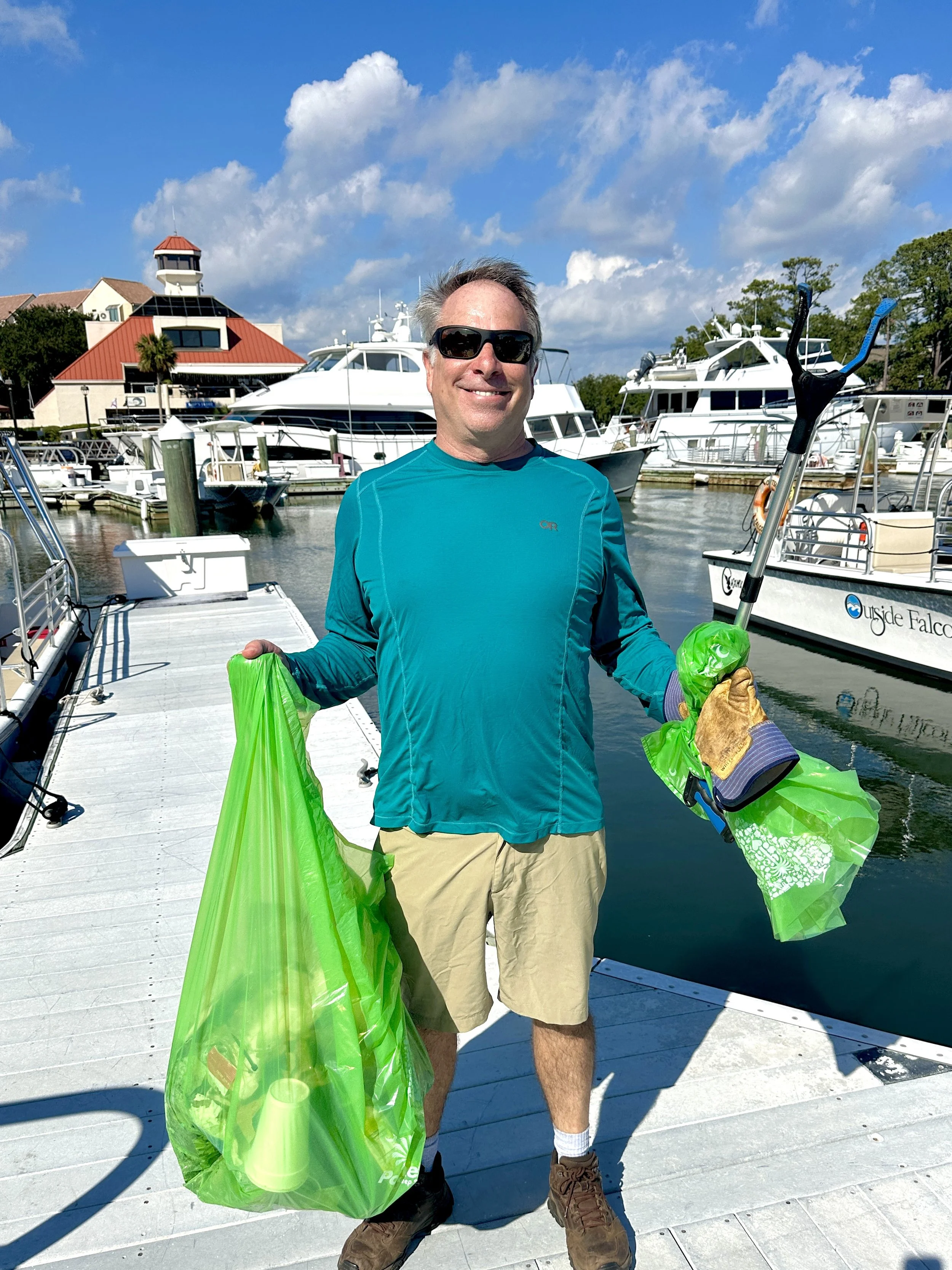 A man wearing sunglasses, a teal long-sleeve shirt, khaki shorts, and hiking boots holding two green plastic bags standing on a dock near boats with a marina and a building with a red roof in the background.