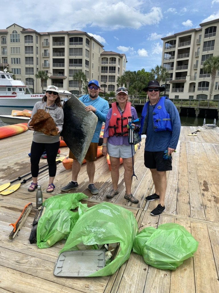 Group of four people on a dock with green trash bags filled with trash and cleaning supplies, holding a large rock and a piece of wood, in front of water and boats, with apartment buildings in the background.
