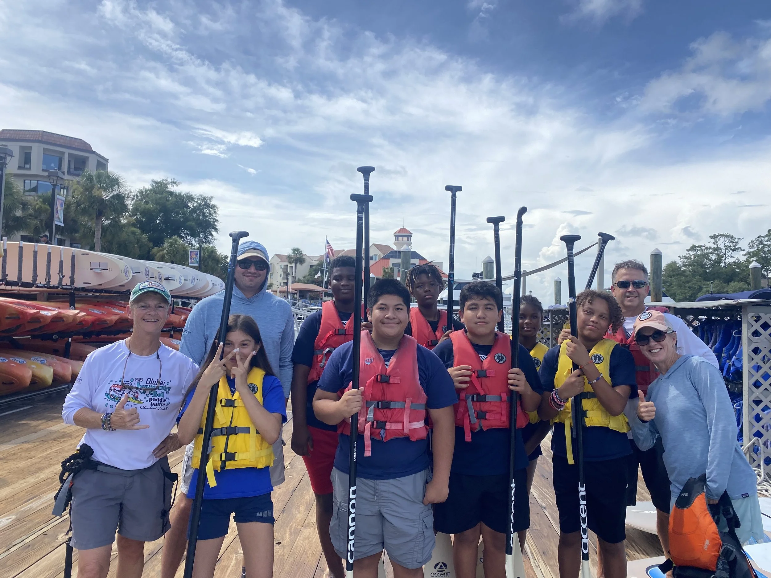 Group of young people and adults gathered on a wooden dock, holding paddles and wearing life jackets, with boats and buildings in the background under a partly cloudy sky.