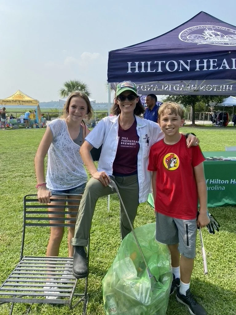 A woman and two children standing outdoors at a community event, smiling for the camera. The woman is holding a trash picker and is dressed casually with sunglasses and a baseball cap. One girl and one boy are standing beside her, with tents and othe