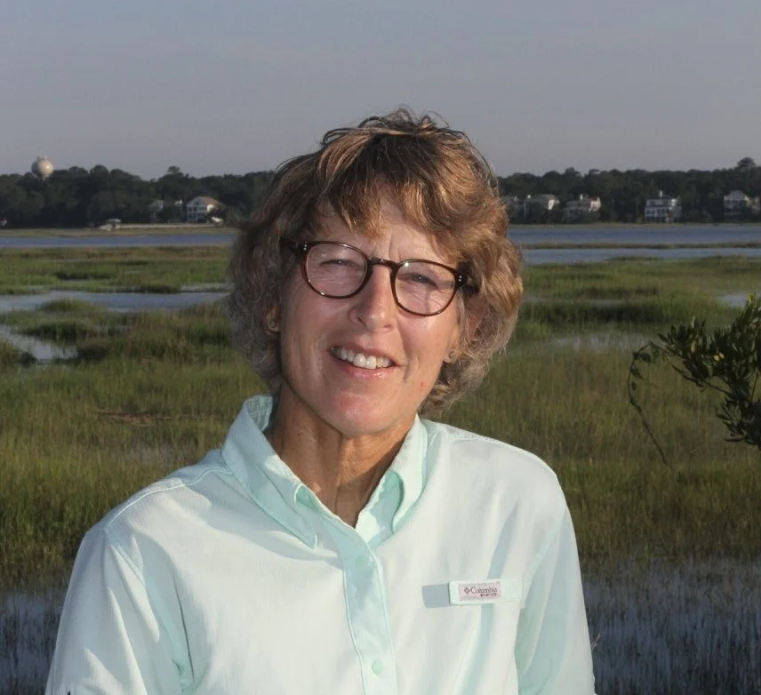 A smiling woman with short curly hair and glasses, wearing a light green collared shirt, standing outdoors near a marshland with a water body and houses in the background.