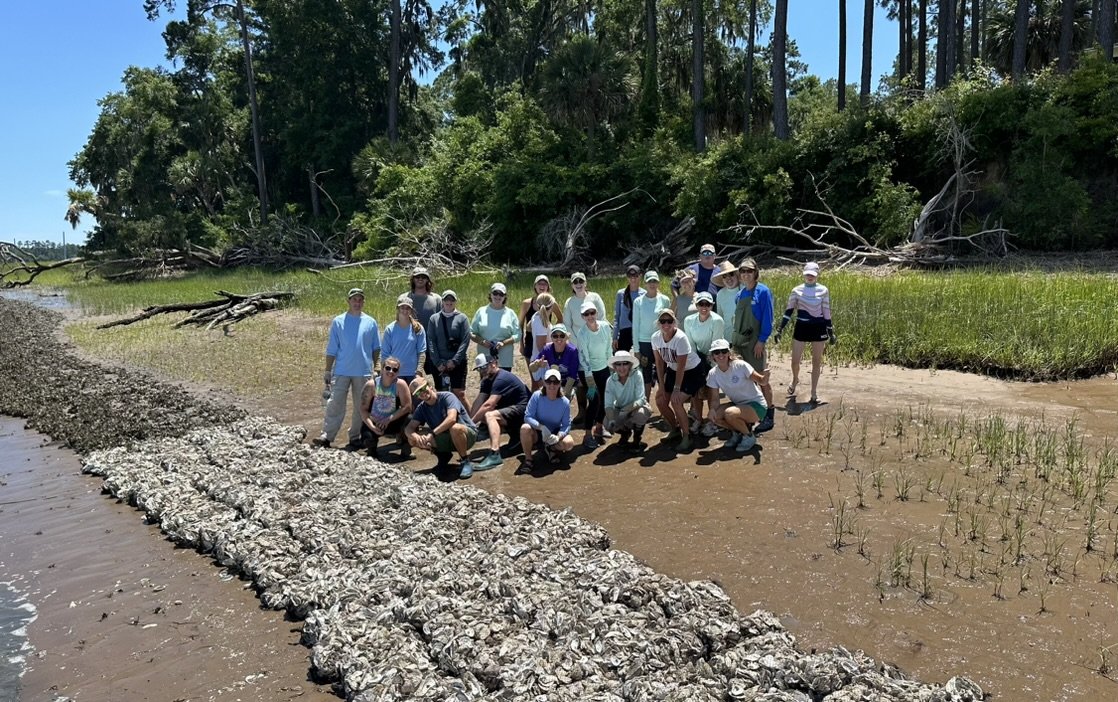 Group of people on a muddy riverbank with grass, trees, and fallen branches in the background on a sunny day.