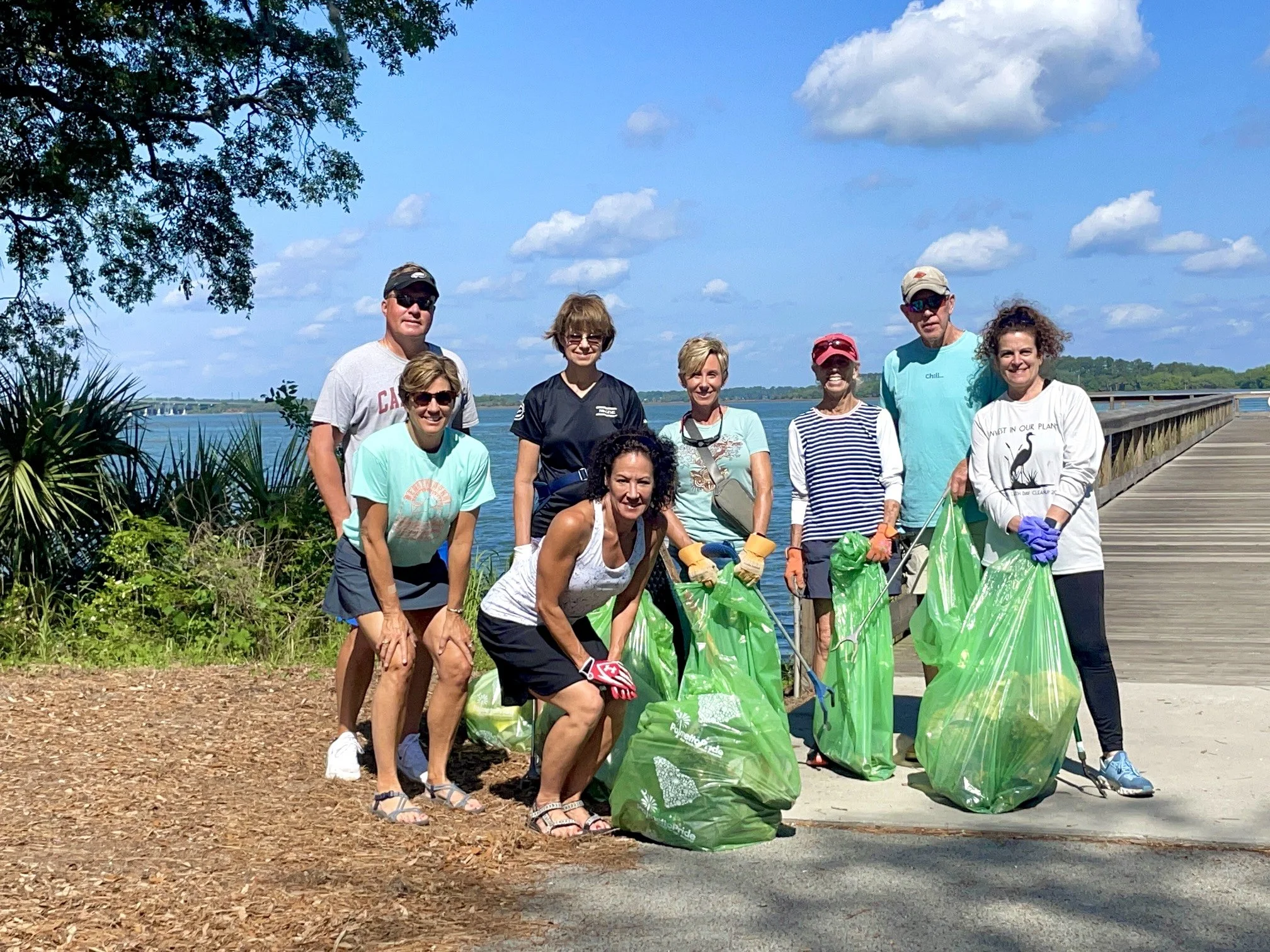 Group of people participating in a community cleanup by a river, holding large green trash bags and cleanup tools, with a wooden walkway and blue sky with clouds in the background.